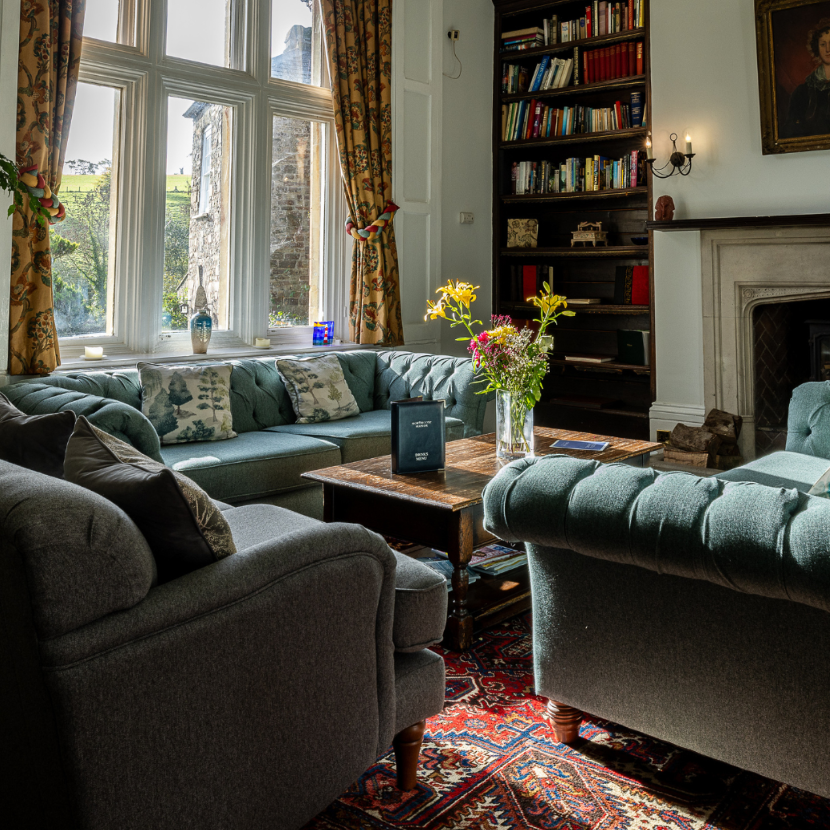Cozy living room with tufted sofas, wooden table, bookshelves, and a large window with floral curtains.