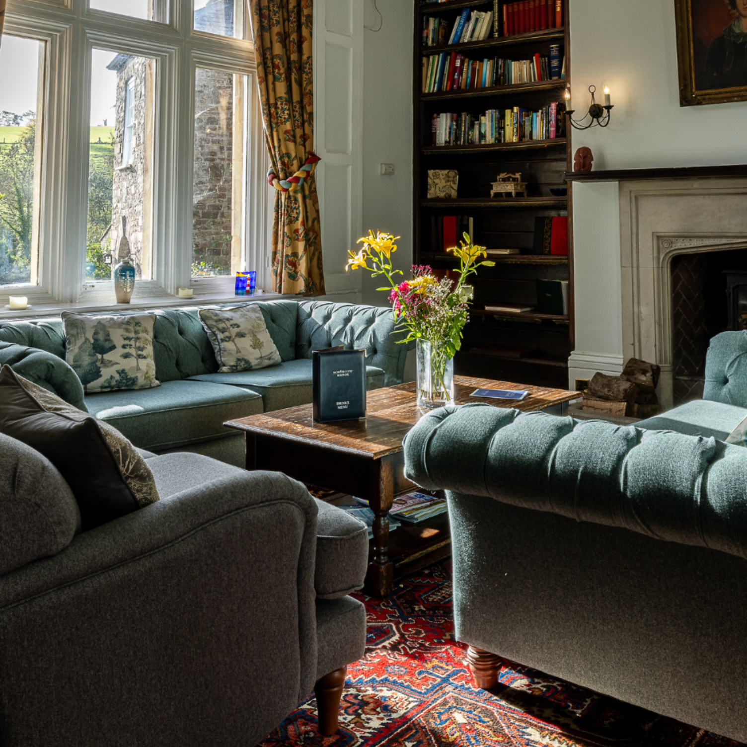 Cozy living room with tufted sofas, wooden table, bookshelves, and a large window with floral curtains.