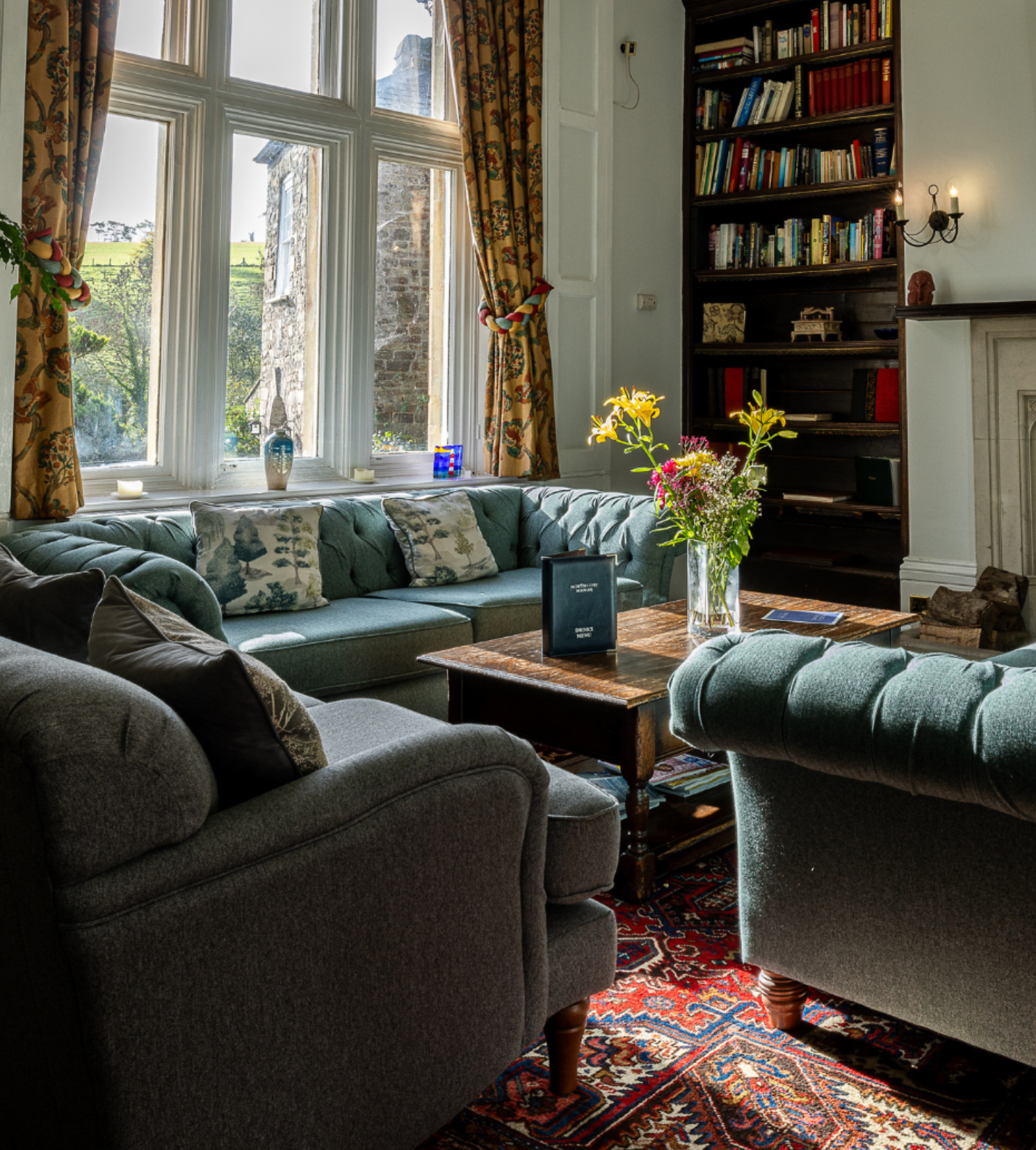 Cozy living room with tufted sofas, wooden table, bookshelves, and a large window with floral curtains.