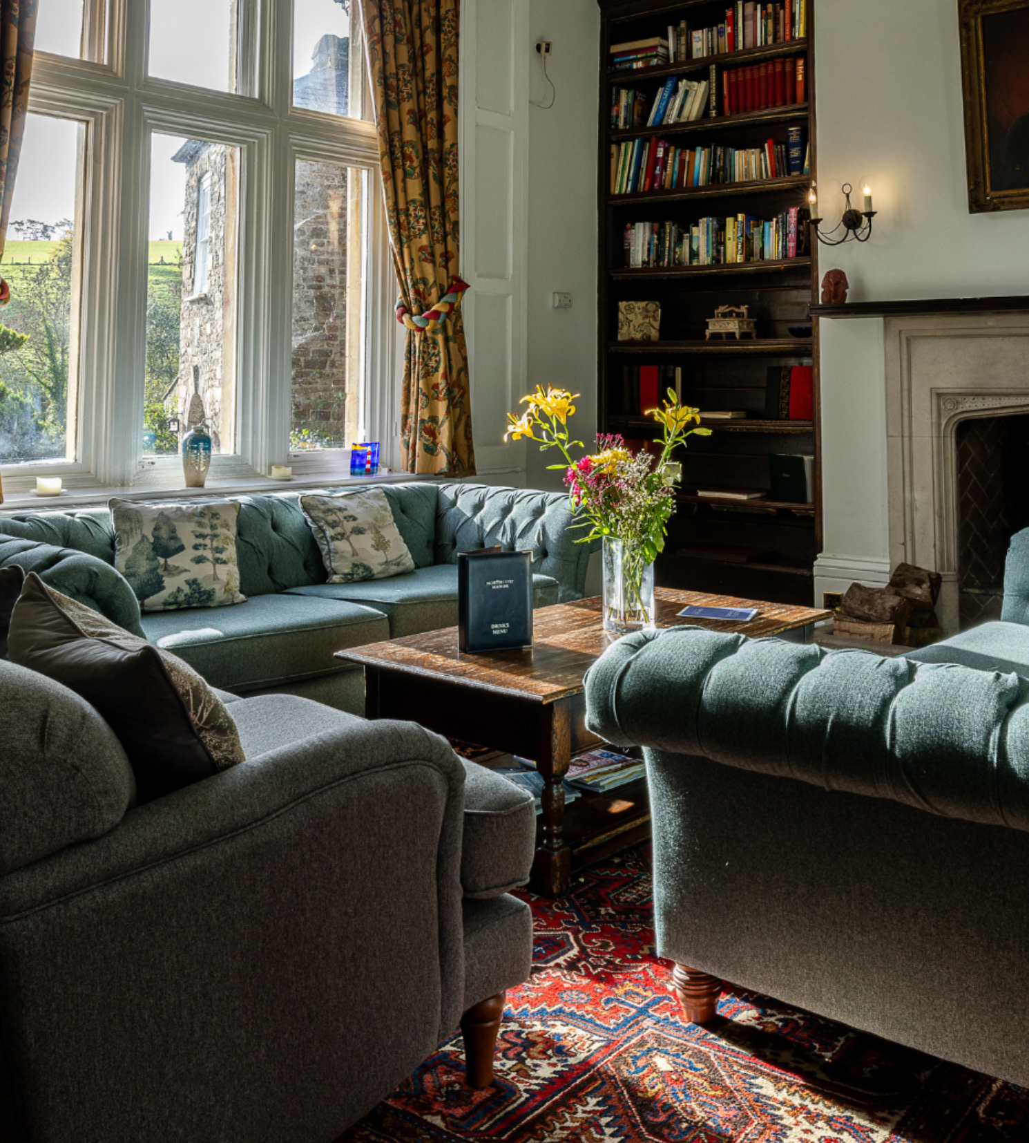 Cozy living room with tufted sofas, wooden table, bookshelves, and a large window with floral curtains.
