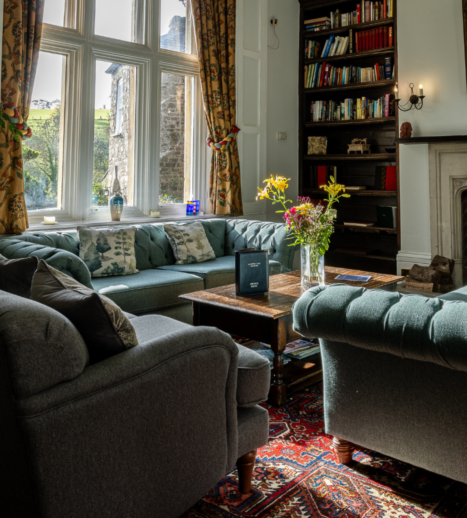 Cozy living room with tufted sofas, wooden table, bookshelves, and a large window with floral curtains.