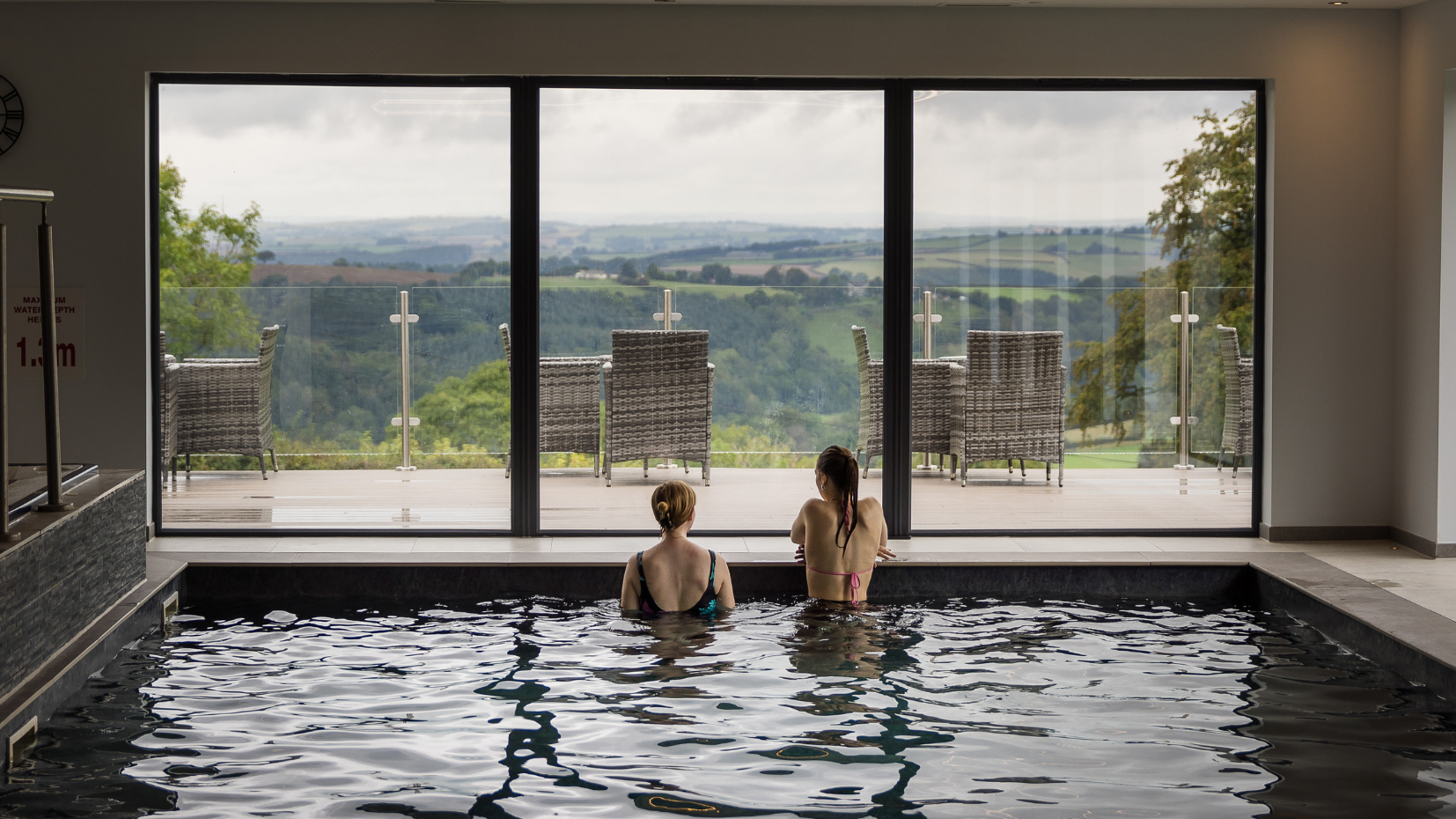 Two people relaxing in an indoor pool with a view of outdoor seating and rolling hills through large glass doors.