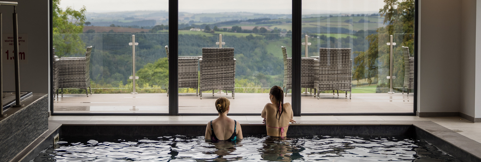 Two people relaxing in an indoor pool with a view of outdoor seating and rolling hills through large glass doors.