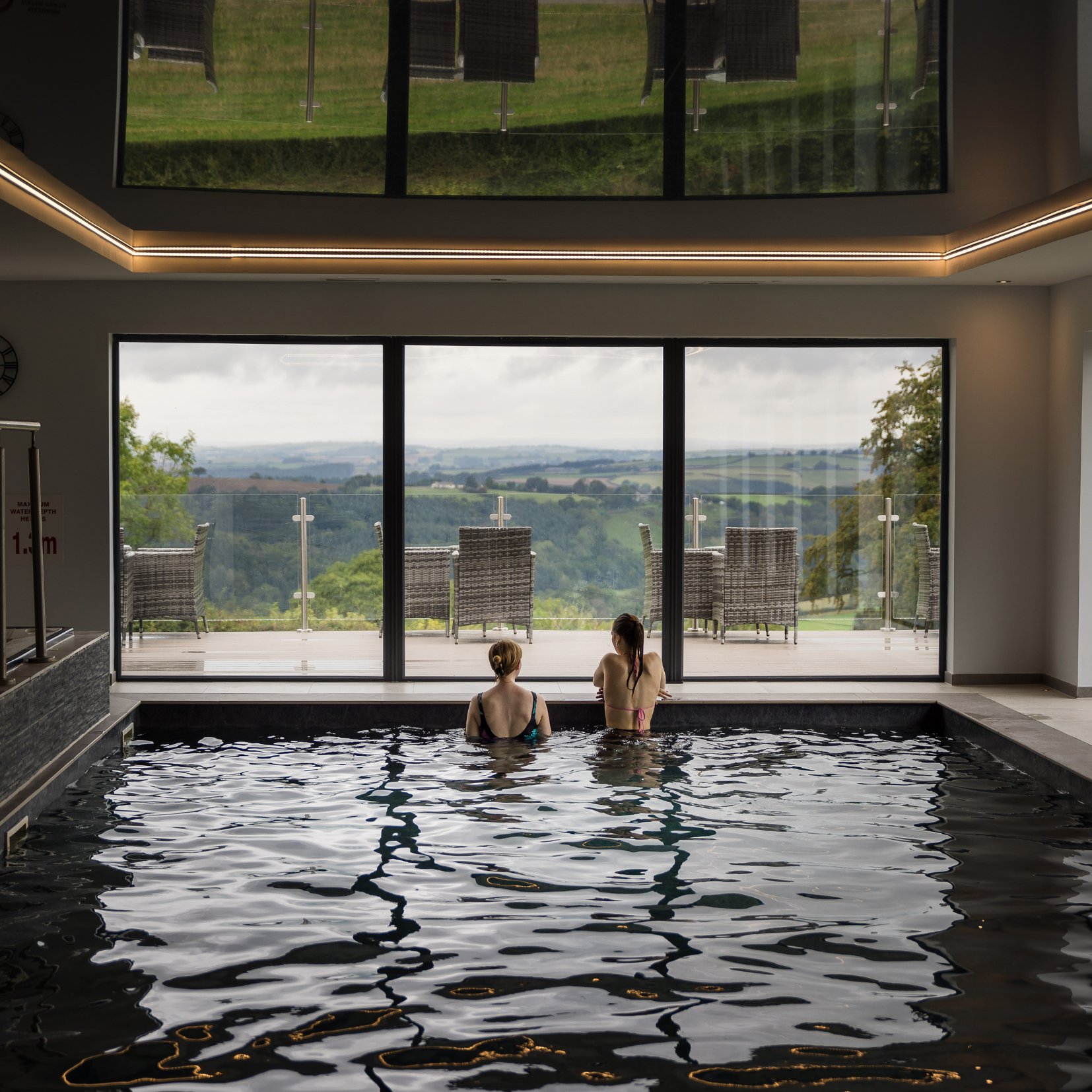 Two people relaxing in an indoor pool with a view of outdoor seating and rolling hills through large glass doors.