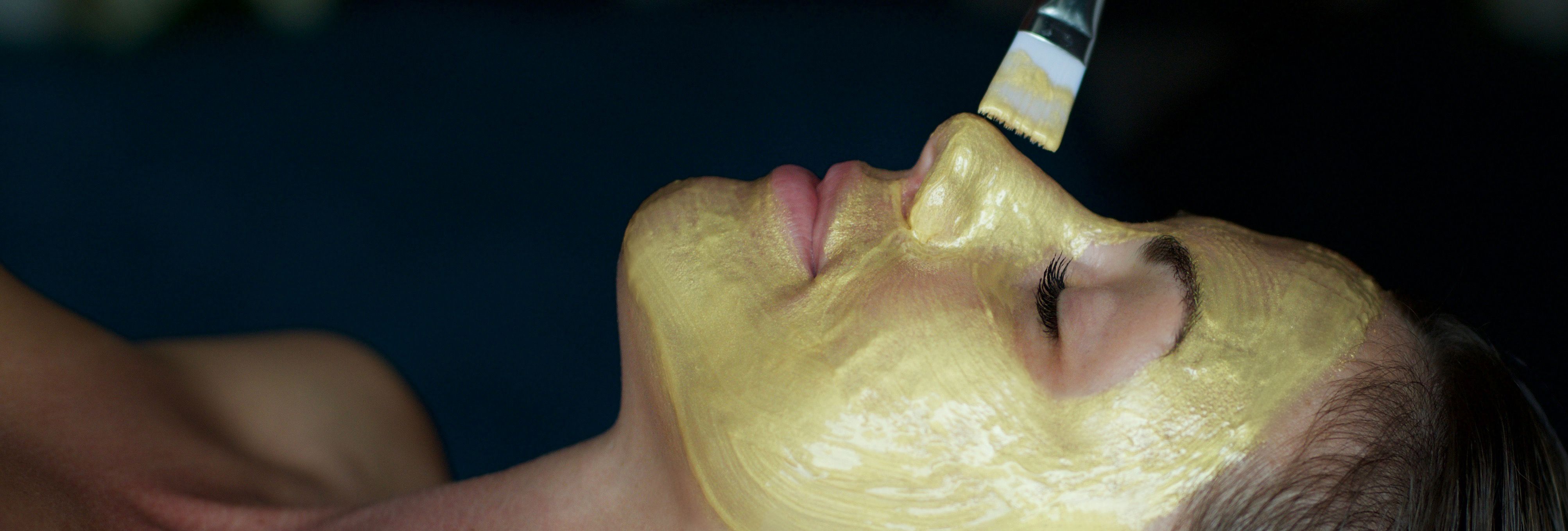 Person receiving a gold facial mask applied with a brush in a spa setting