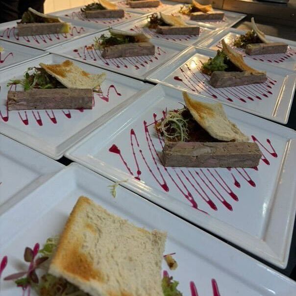 Multiple square plates with slices of toasted bread, pâté, greens, and a pink sauce drizzle arranged in a kitchen.