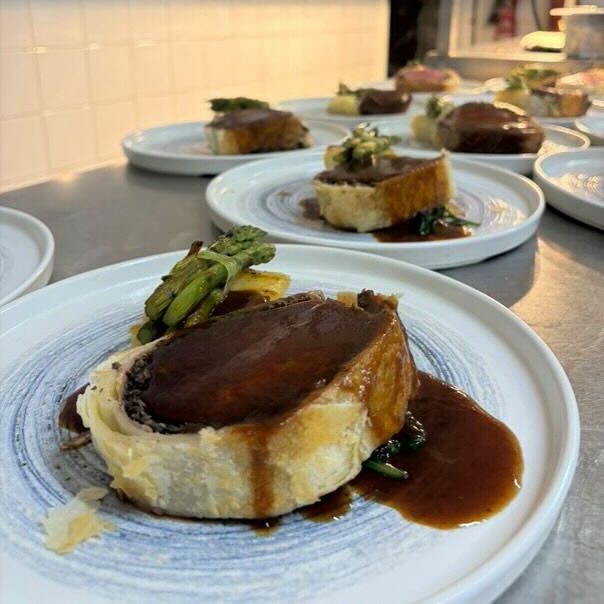 Plated servings of a meat dish with pastry and brown sauce, garnished with vegetables, arranged on a kitchen counter.