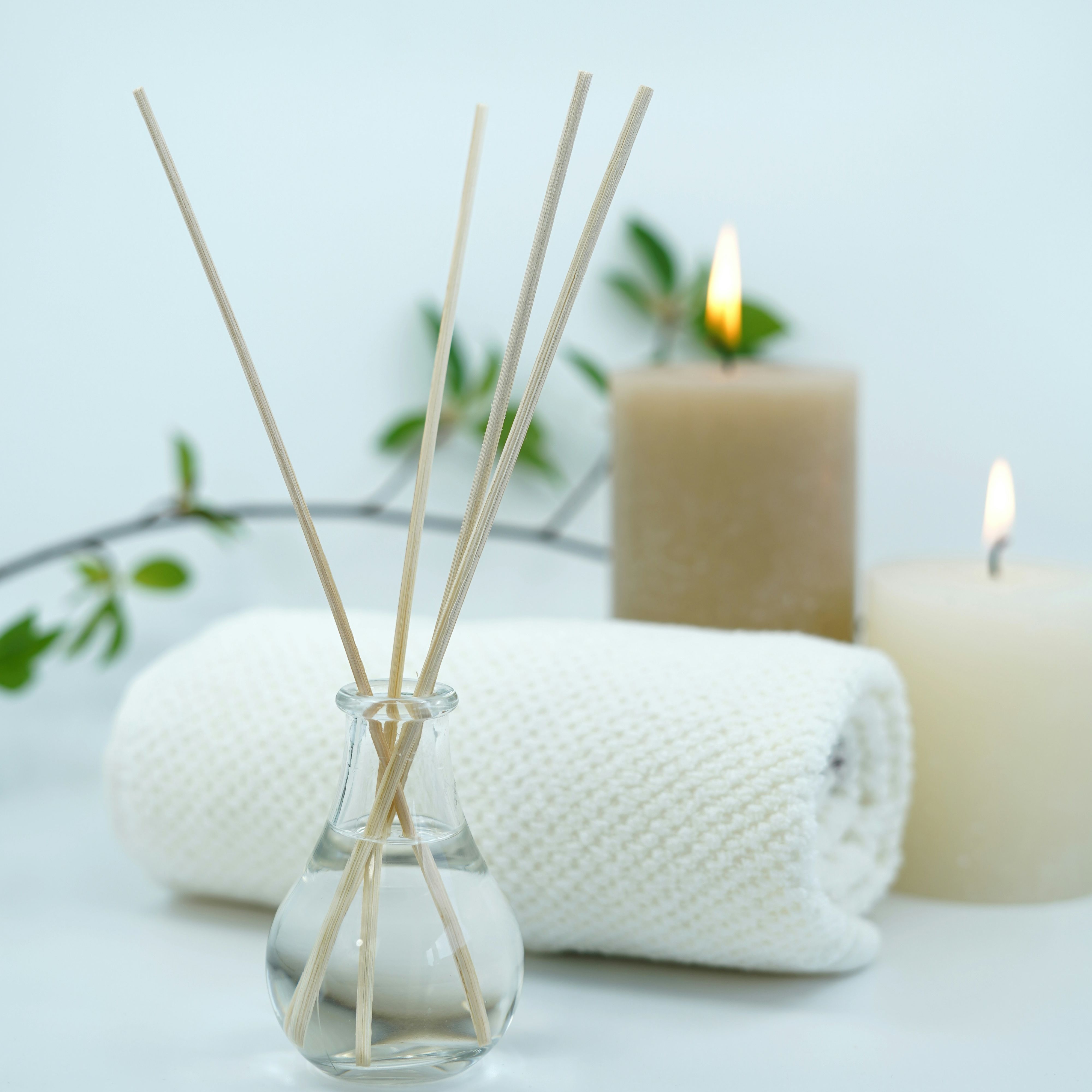 Aromatic reed diffuser, rolled towel, and lit candles with green leaves in the background.