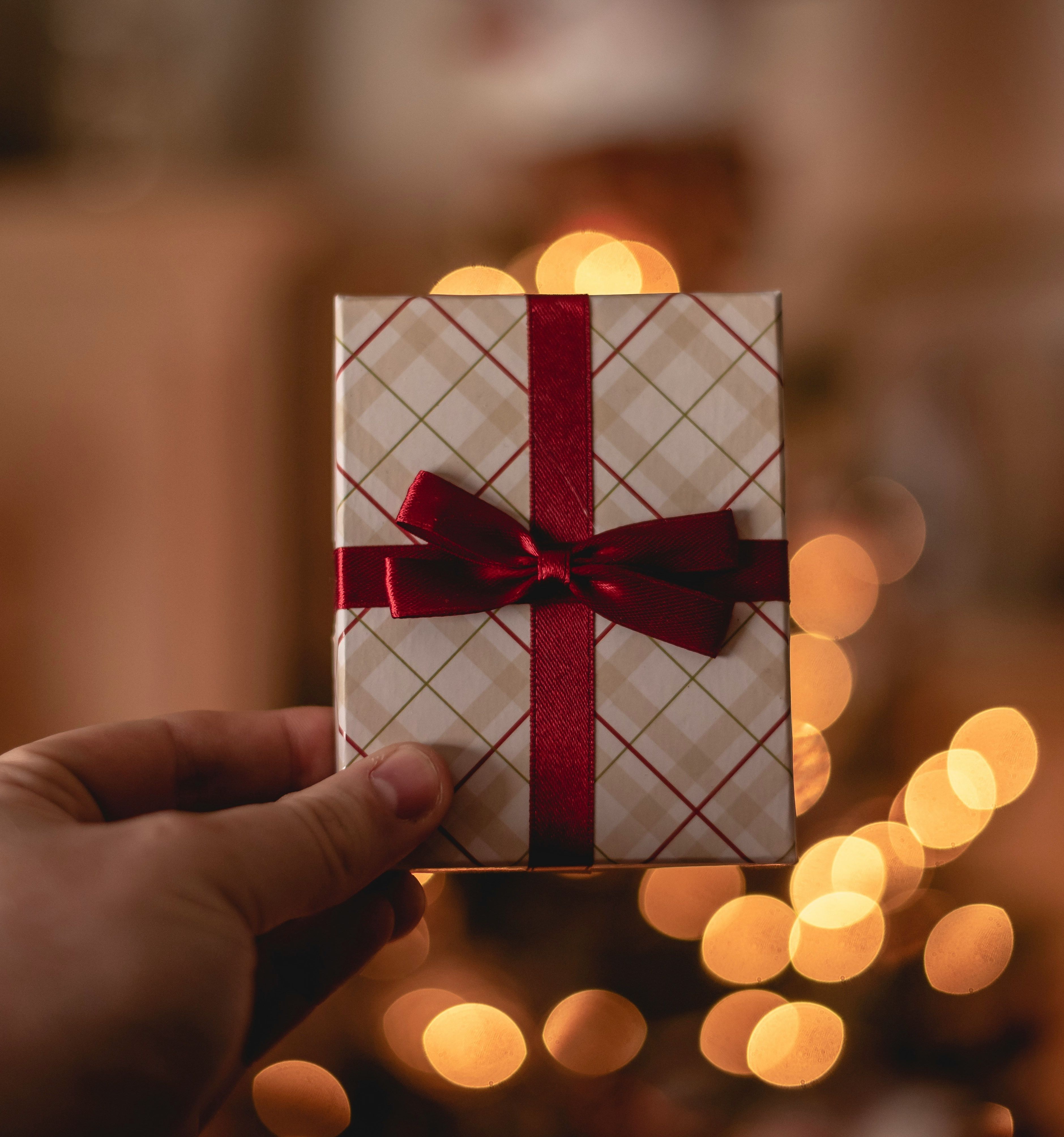 Hand holding a small gift wrapped with red ribbon in front of warm holiday lights
