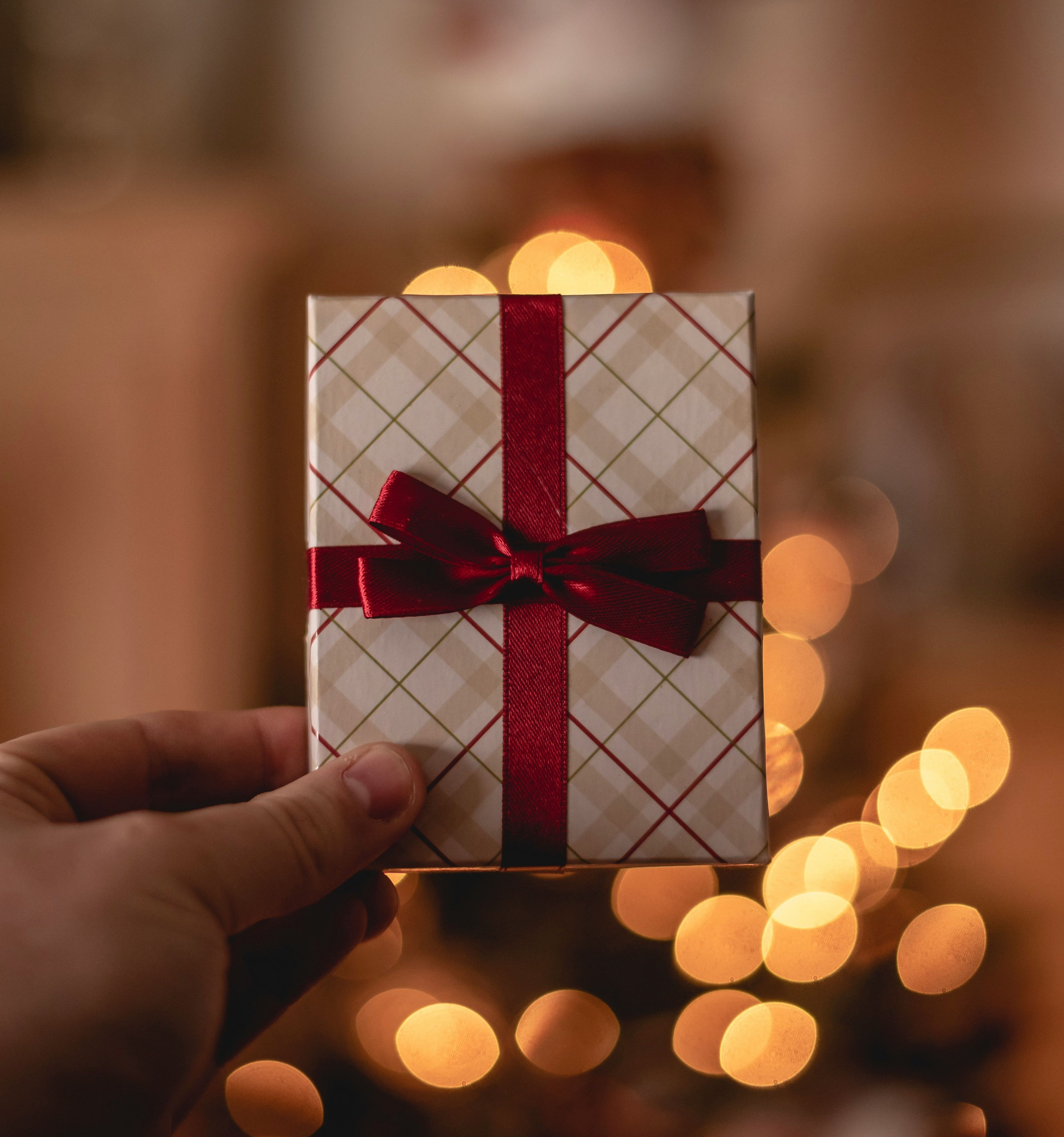 Hand holding a small gift wrapped with red ribbon in front of warm holiday lights