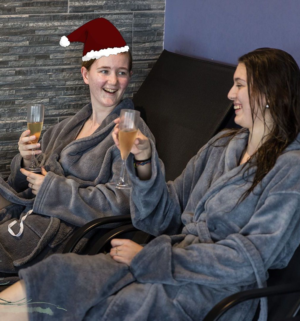 Two people in spa robes relaxing on lounge chairs, holding drinks, with festive holiday decorations including a Santa hat, cartoon Christmas tree, and snowflakes.