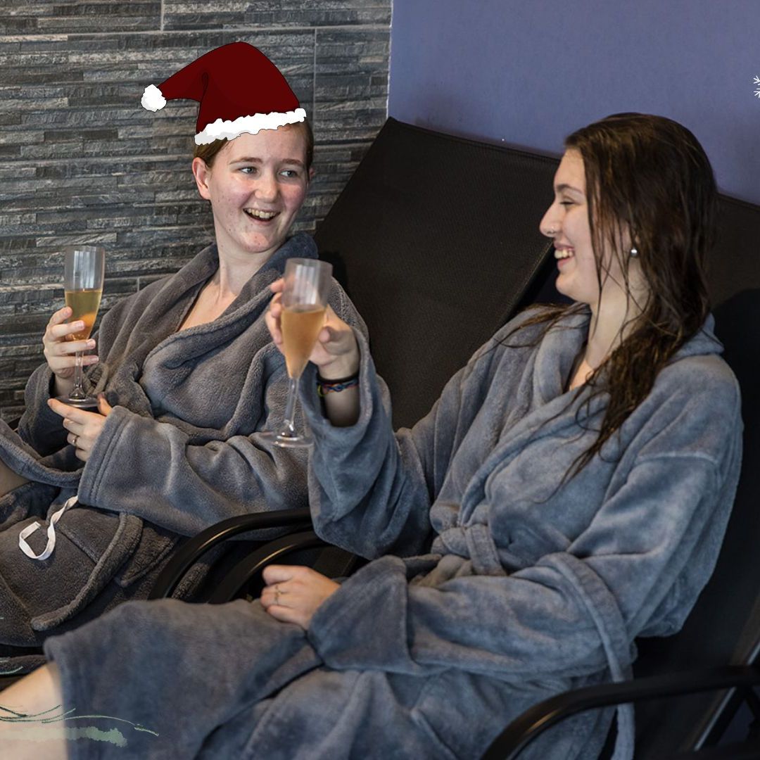 Two people in spa robes relaxing on lounge chairs, holding drinks, with festive holiday decorations including a Santa hat, cartoon Christmas tree, and snowflakes.