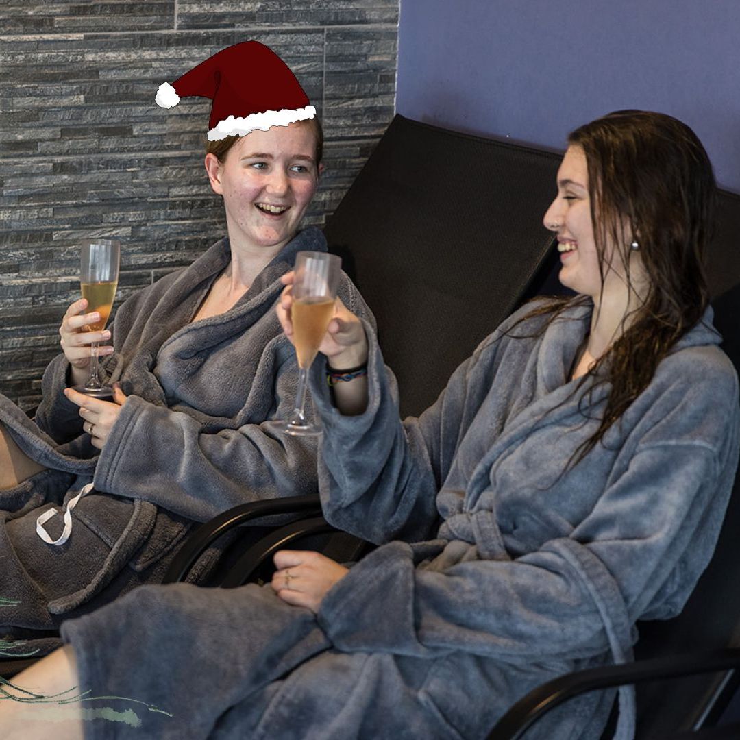 Two people in spa robes relaxing on lounge chairs, holding drinks, with festive holiday decorations including a Santa hat, cartoon Christmas tree, and snowflakes.