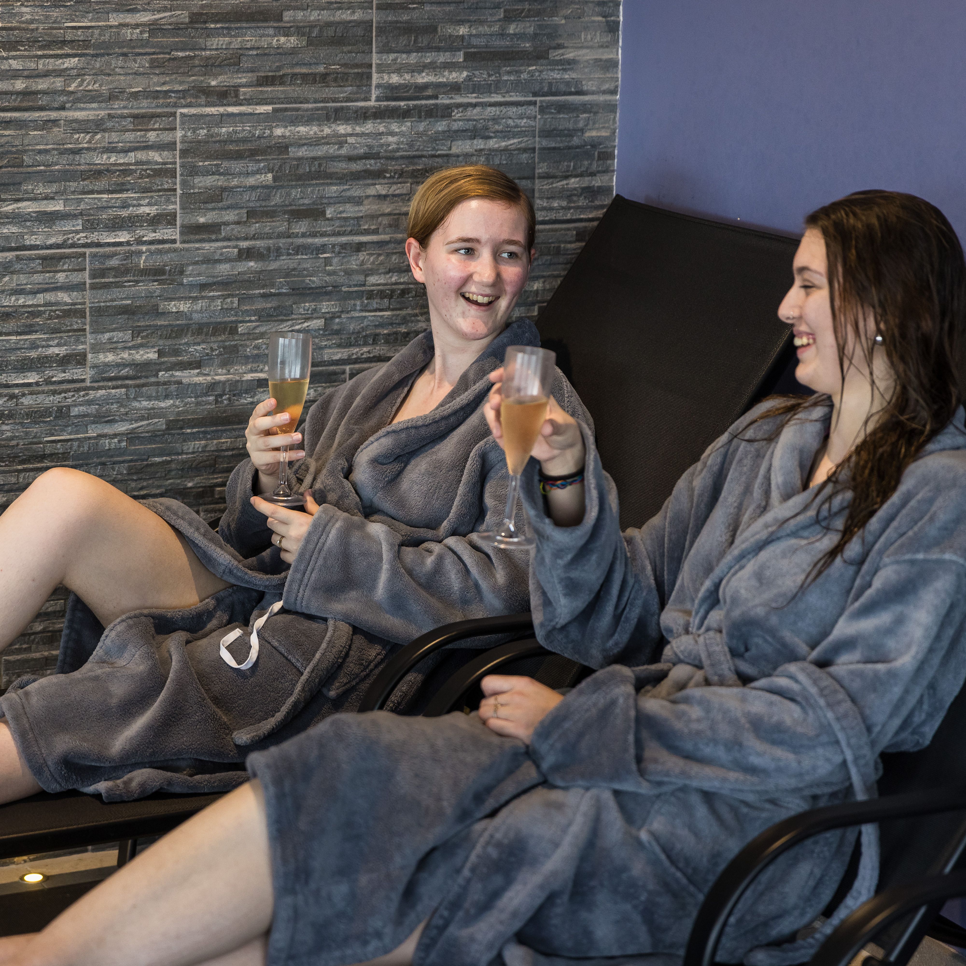 Two people in grey robes enjoying drinks and relaxing on lounge chairs at a spa.