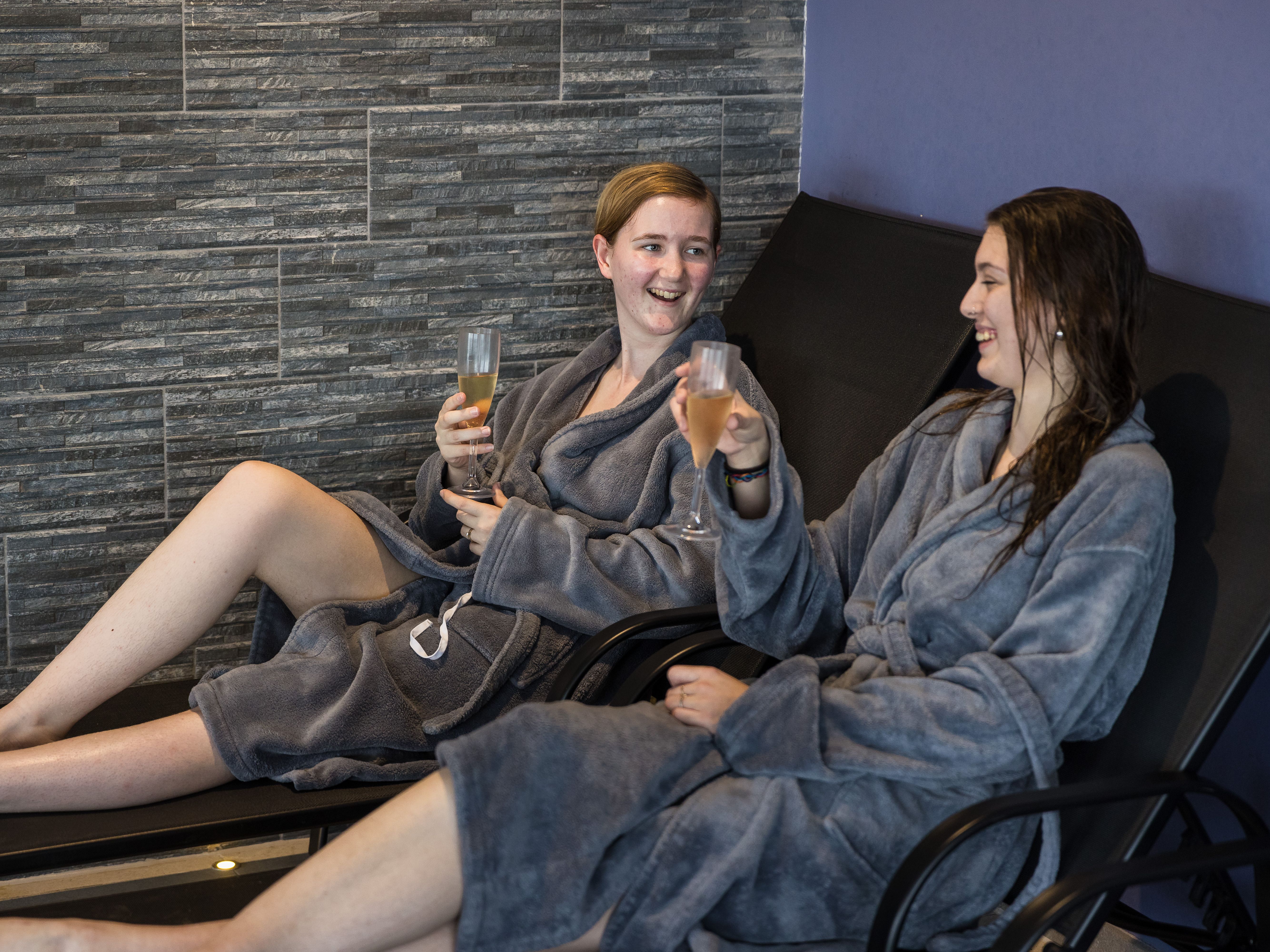 Two people in grey robes enjoying drinks and relaxing on lounge chairs at a spa.