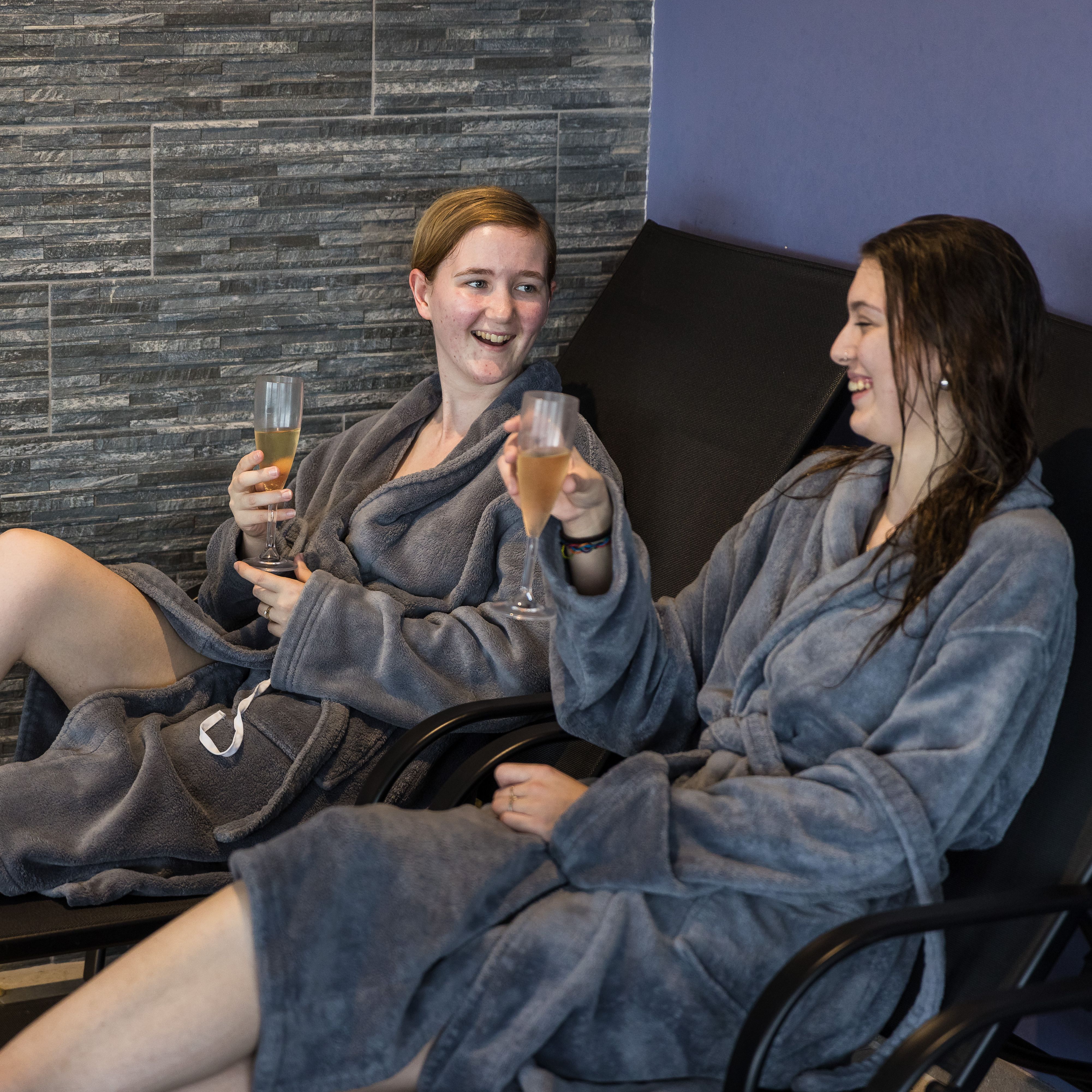 Two people in grey robes enjoying drinks and relaxing on lounge chairs at a spa.