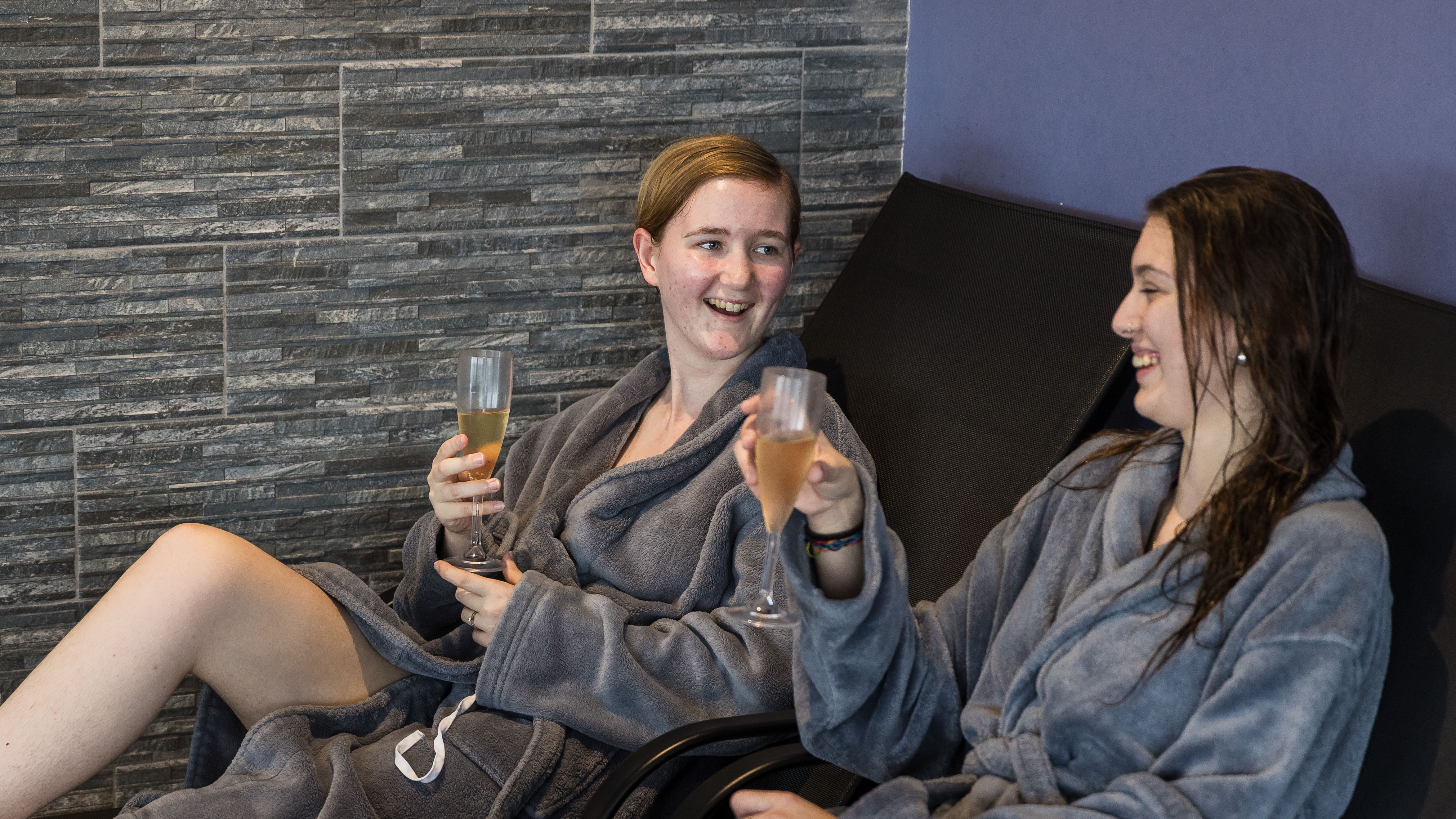 Two people in grey robes enjoying drinks and relaxing on lounge chairs at a spa.