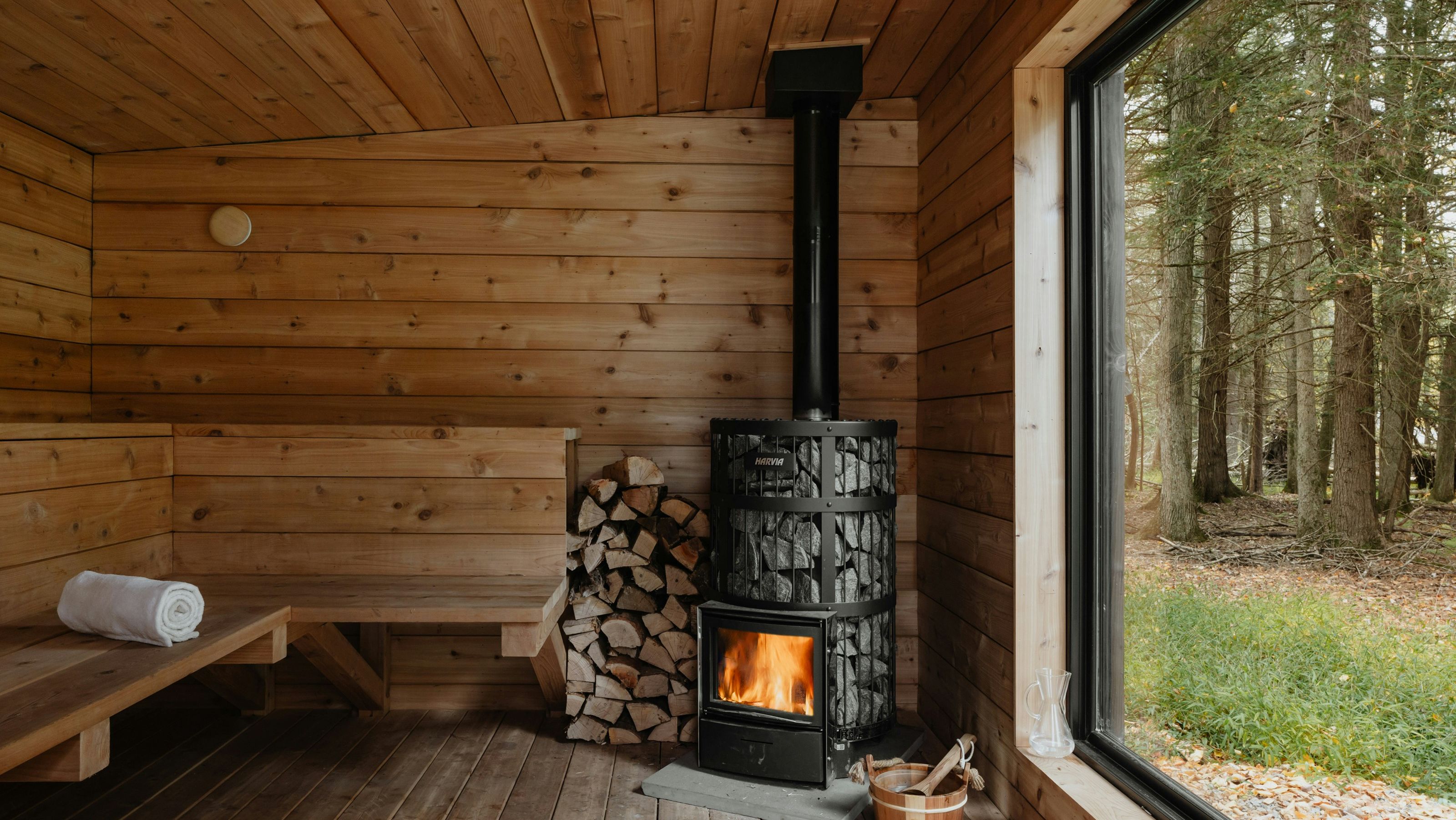 Interior of a wooden sauna with a wood-burning stove, stack of firewood, and a large window overlooking a forest.