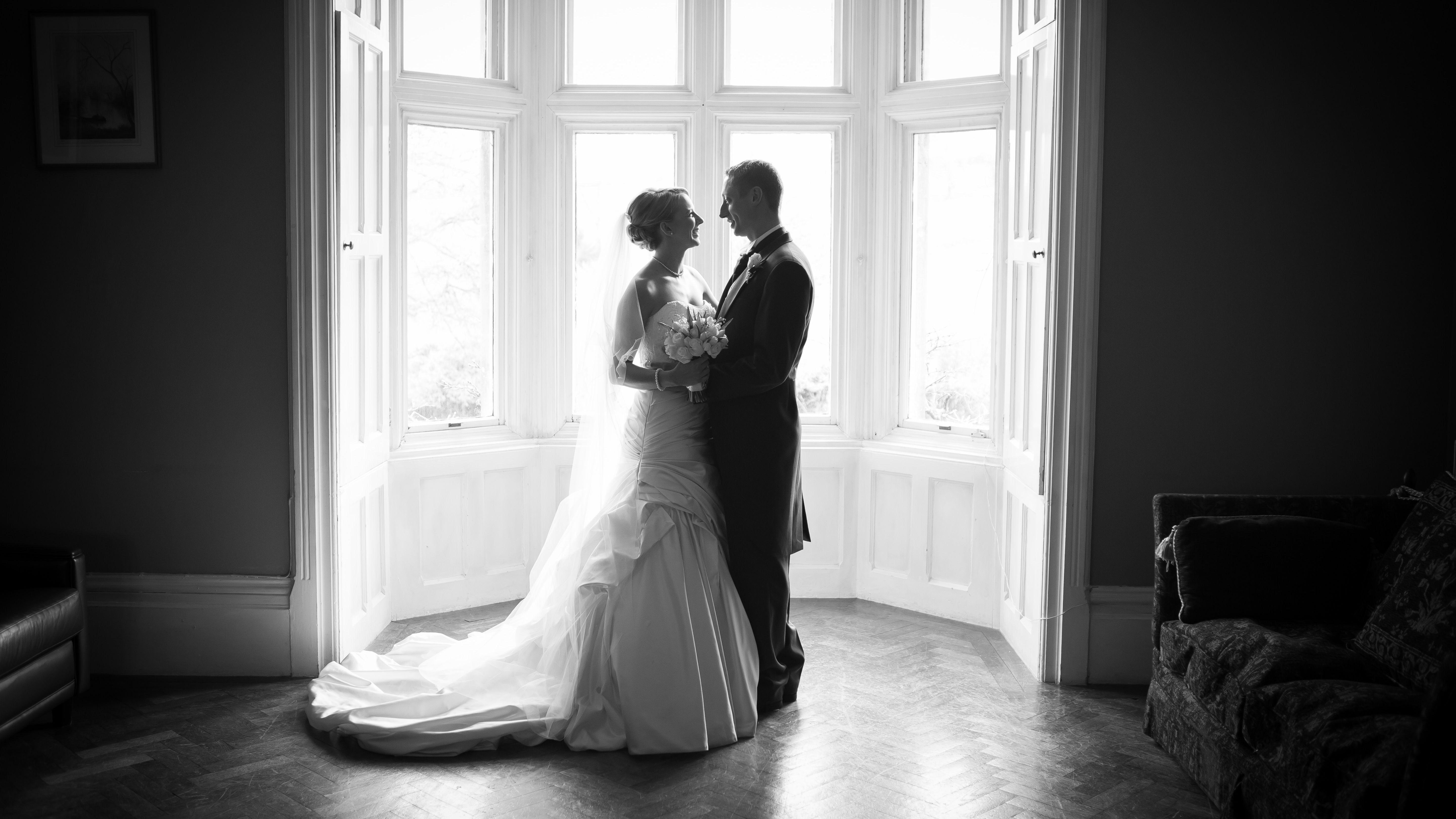 Bride and groom standing together by a large window in a black and white photograph