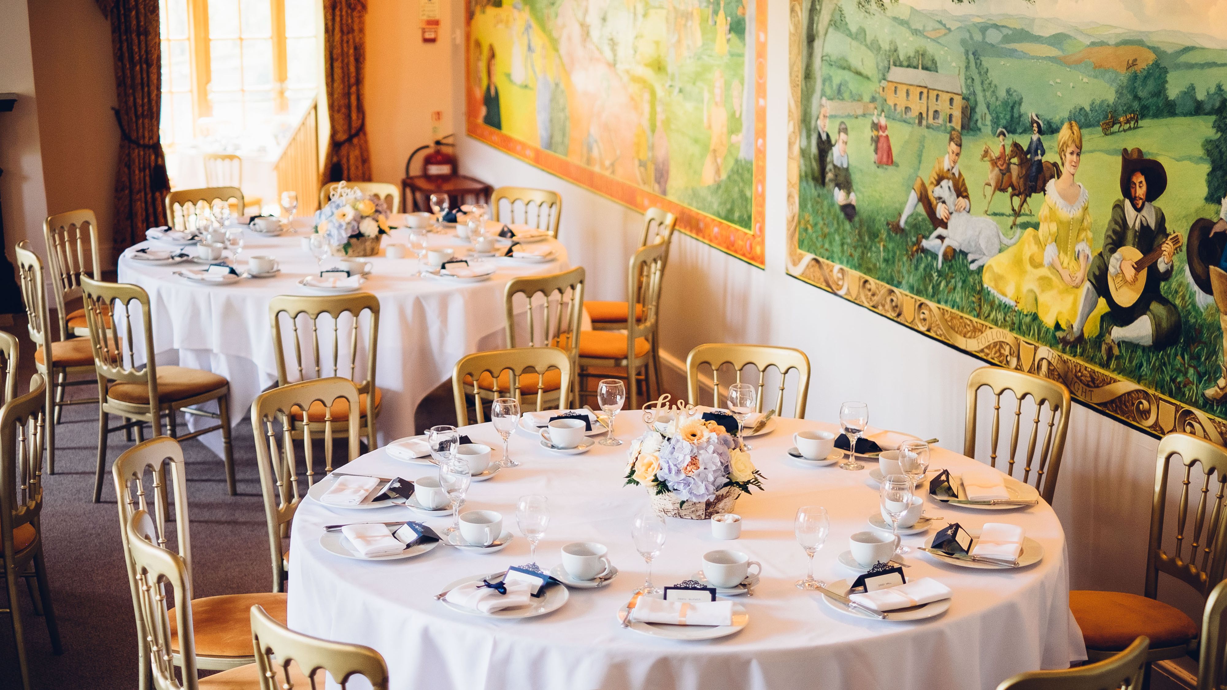 Elegant wedding reception area with round tables set for guests, featuring white tablecloths, gold chairs, floral centerpieces, and a colorful mural on the wall.