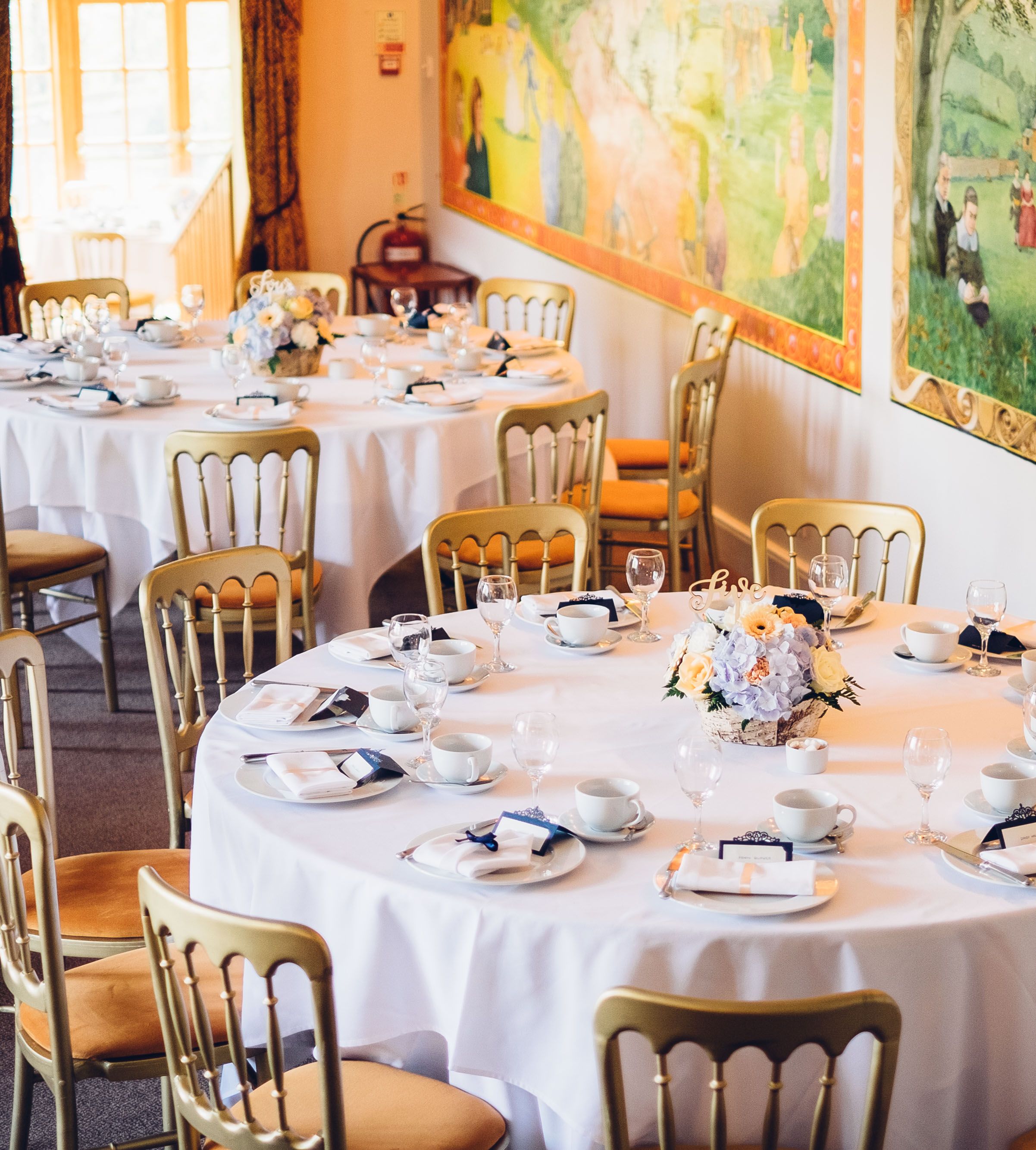 Elegant wedding reception area with round tables set for guests, featuring white tablecloths, gold chairs, floral centerpieces, and a colorful mural on the wall.