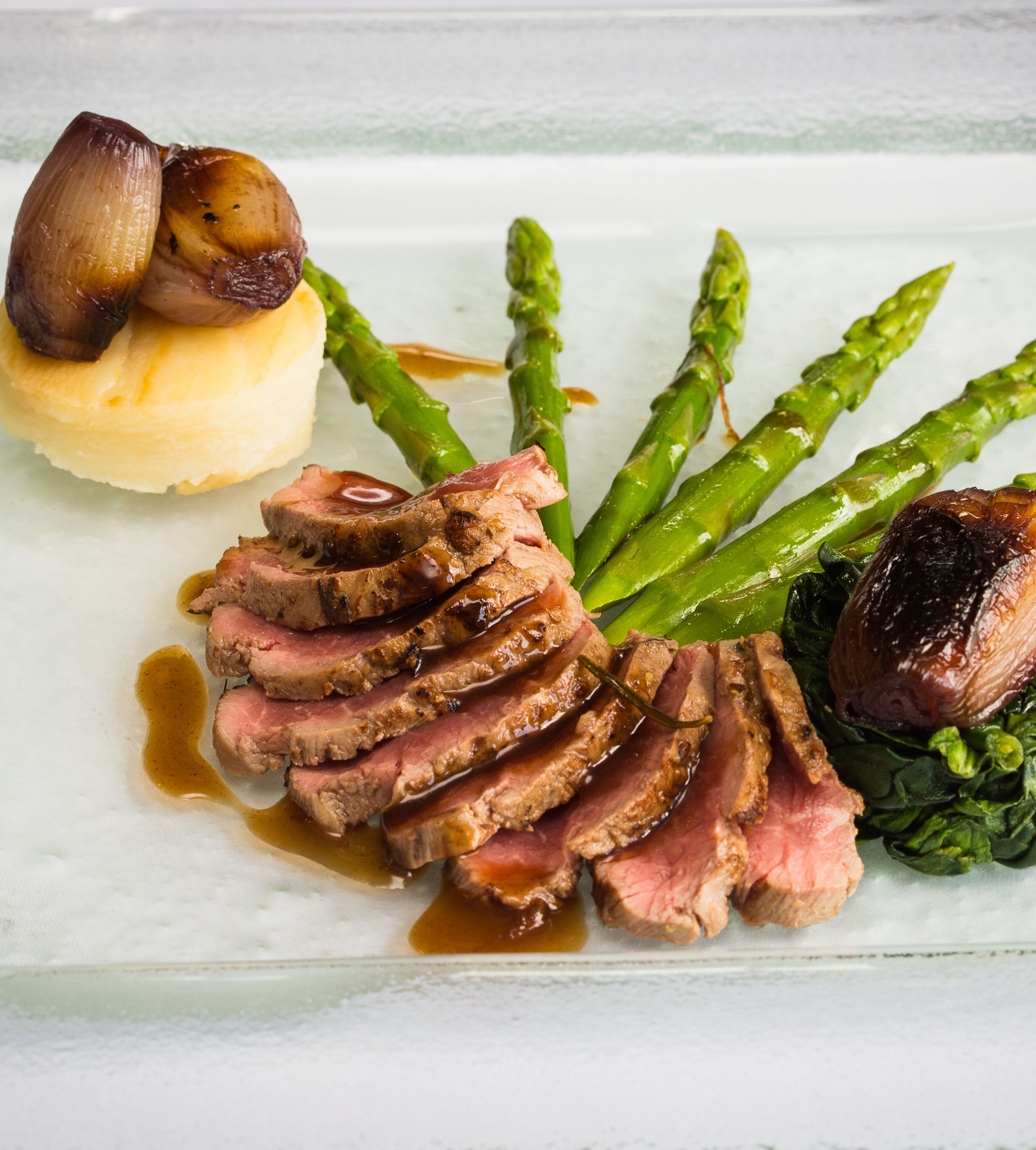 Gourmet plated dish with sliced beef, mashed potatoes, caramelized shallots, asparagus, and spinach