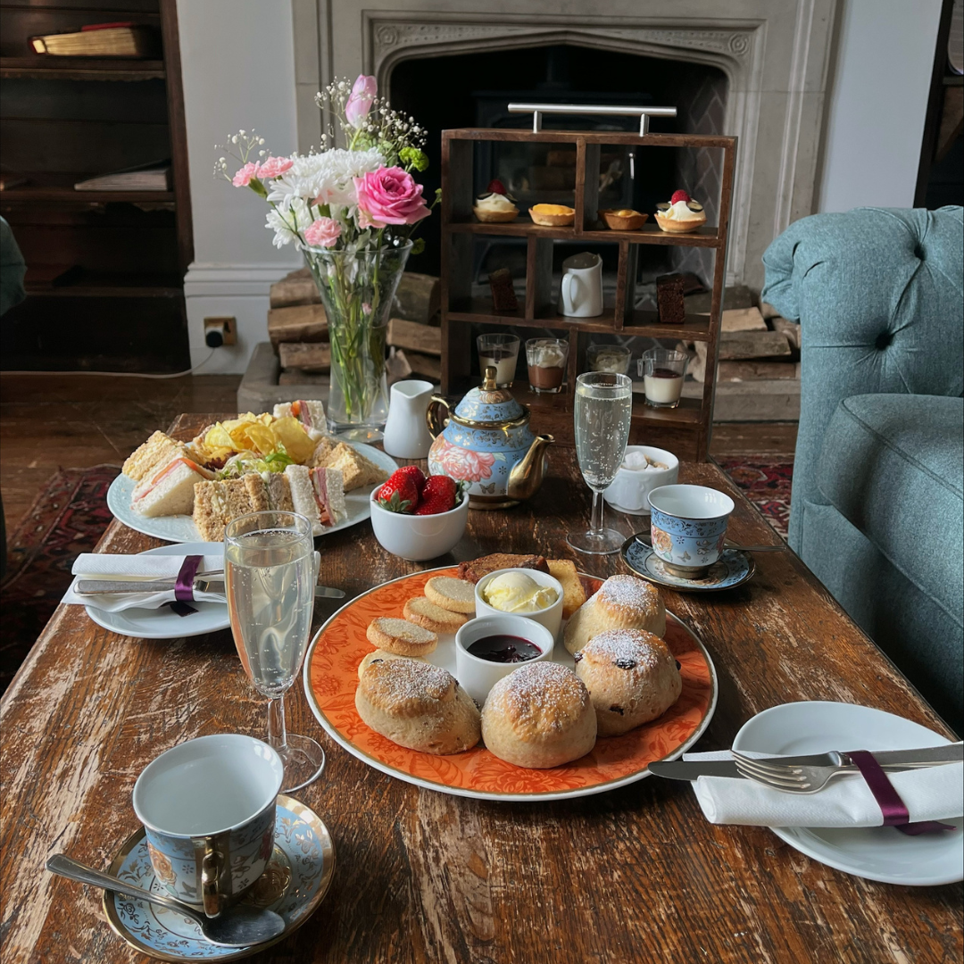 A table set for afternoon tea with scones, sandwiches, pastries, tea cups, and a teapot, next to a vase of flowers in front of a fireplace.