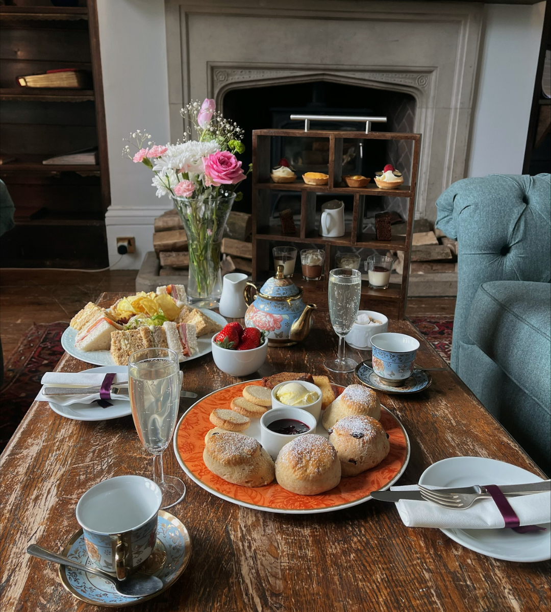 A table set for afternoon tea with scones, sandwiches, pastries, tea cups, and a teapot, next to a vase of flowers in front of a fireplace.