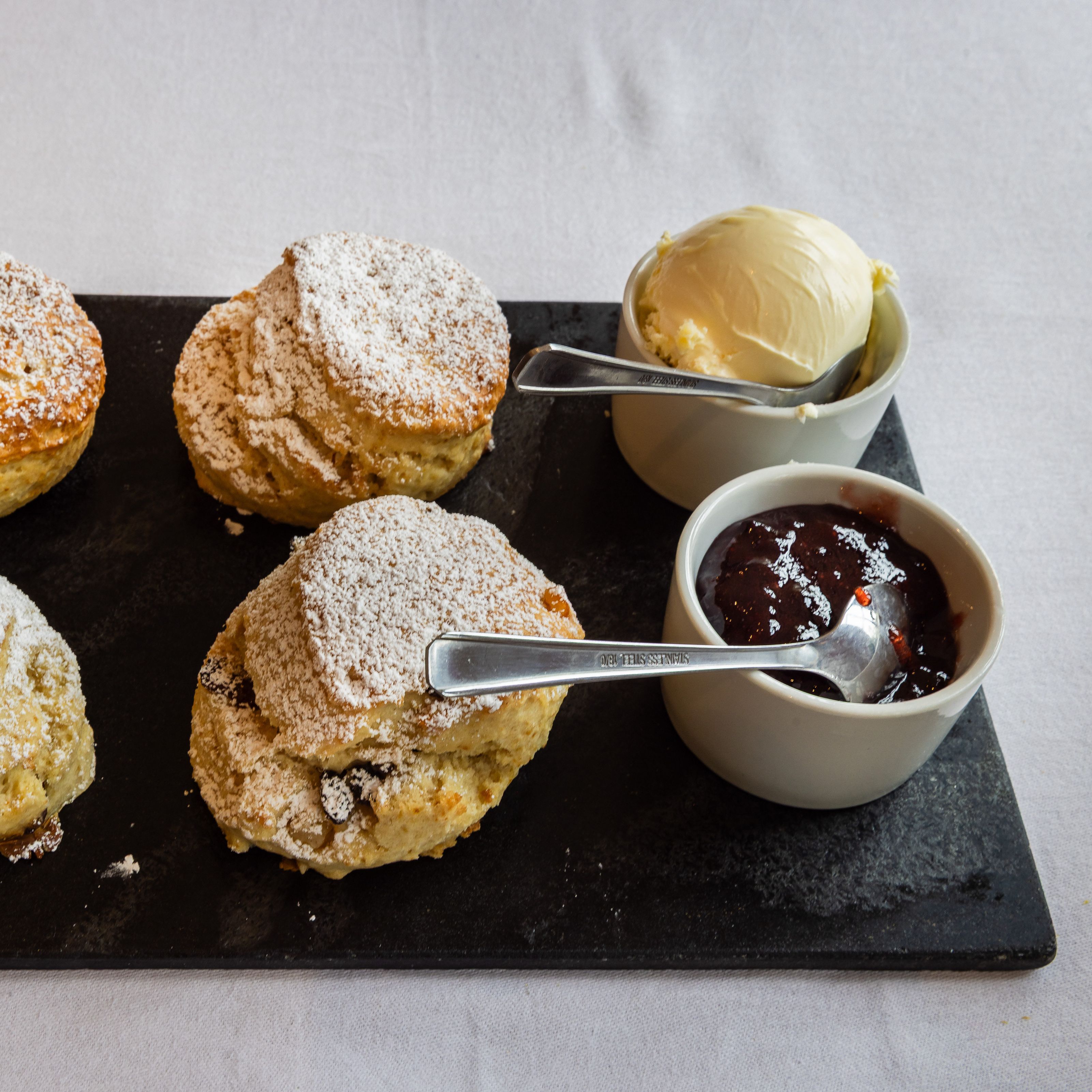 Four scones dusted with powdered sugar served on a slate platter with clotted cream and strawberry jam in small bowls.