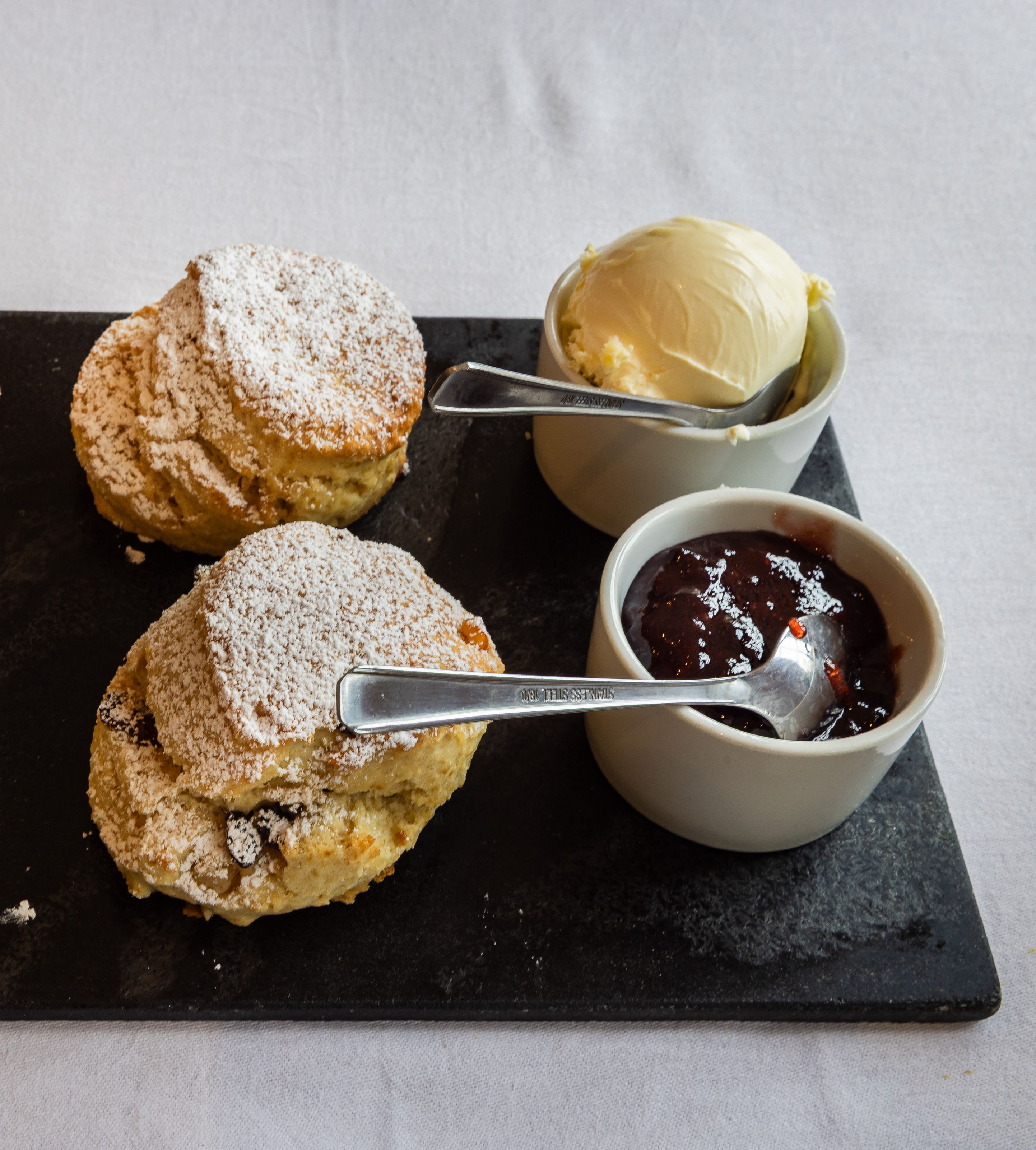 Four scones dusted with powdered sugar served on a slate platter with clotted cream and strawberry jam in small bowls.
