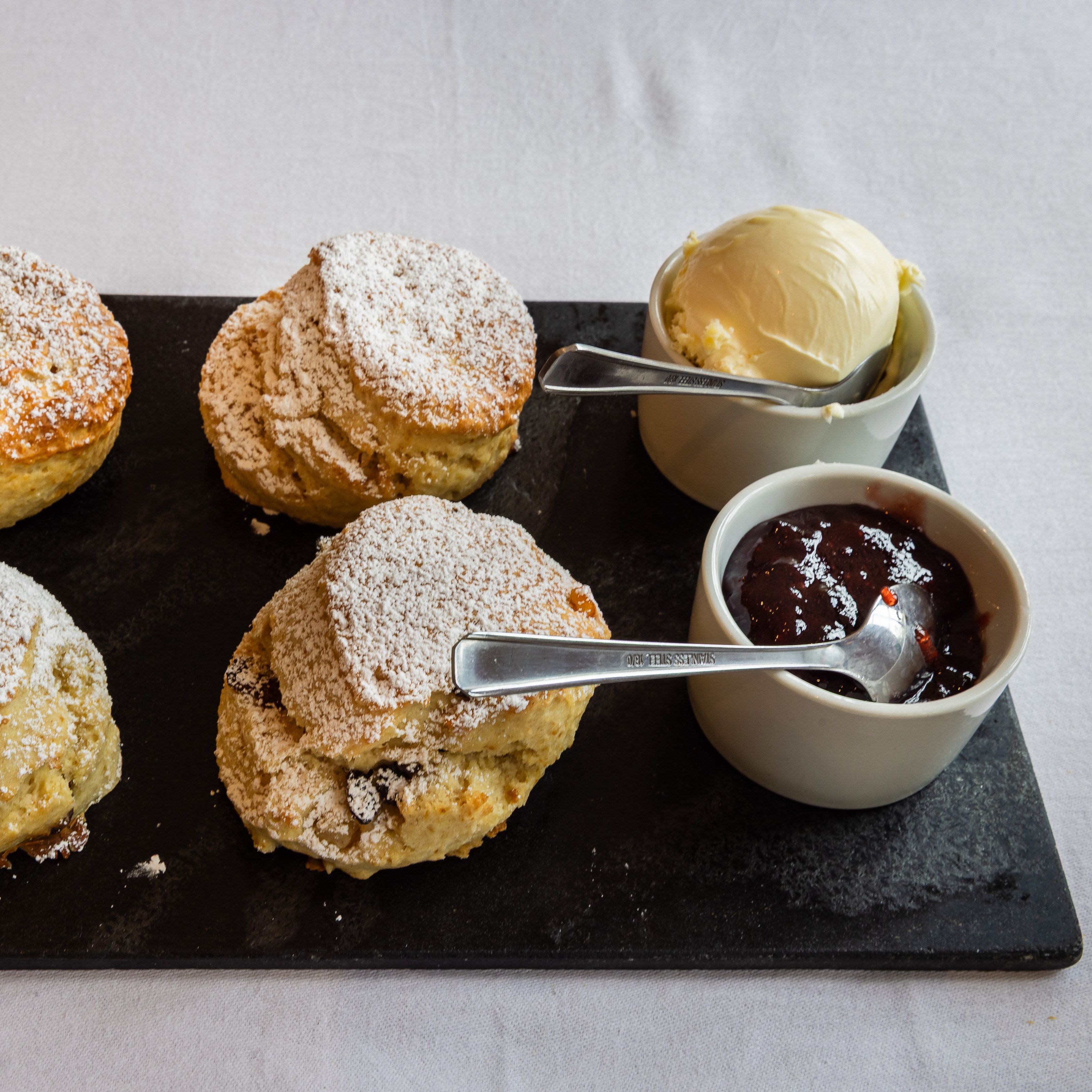 Four scones dusted with powdered sugar served on a slate platter with clotted cream and strawberry jam in small bowls.
