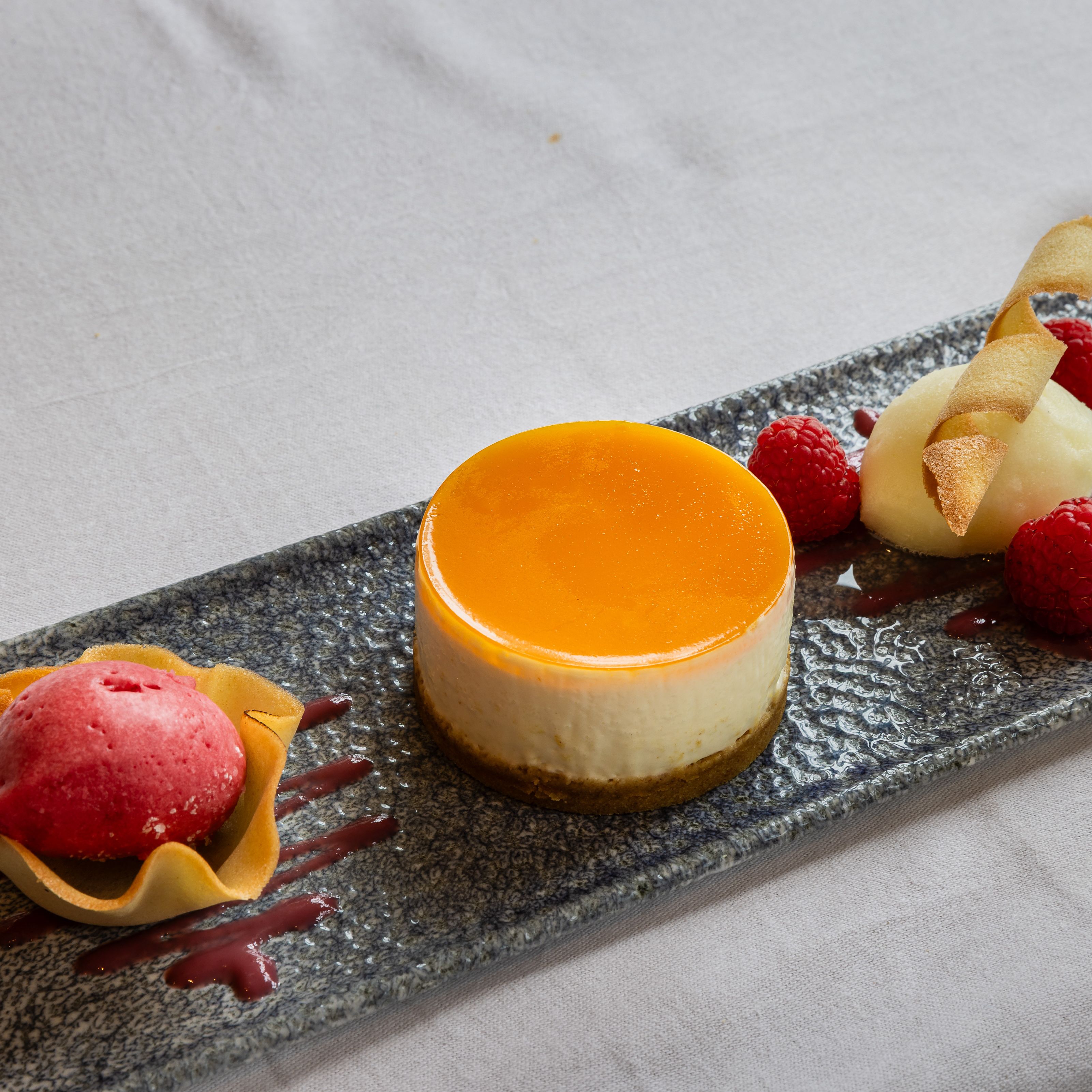 Elegant plated dessert featuring a round cheesecake with orange glaze, raspberry sorbet in a tuile cup, fresh raspberries, and a quenelle of white sorbet garnished with a crisp wafer.