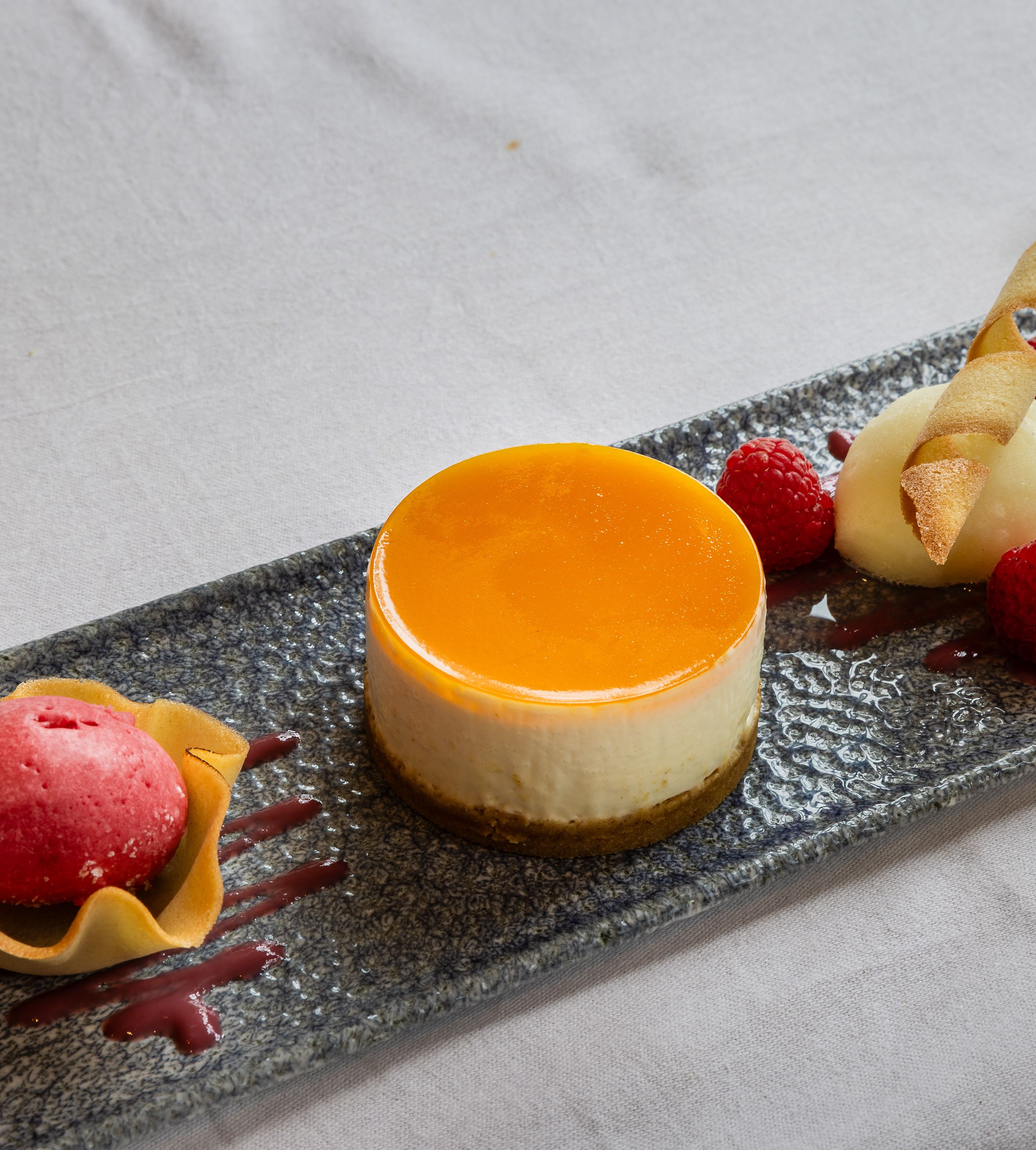 Elegant plated dessert featuring a round cheesecake with orange glaze, raspberry sorbet in a tuile cup, fresh raspberries, and a quenelle of white sorbet garnished with a crisp wafer.