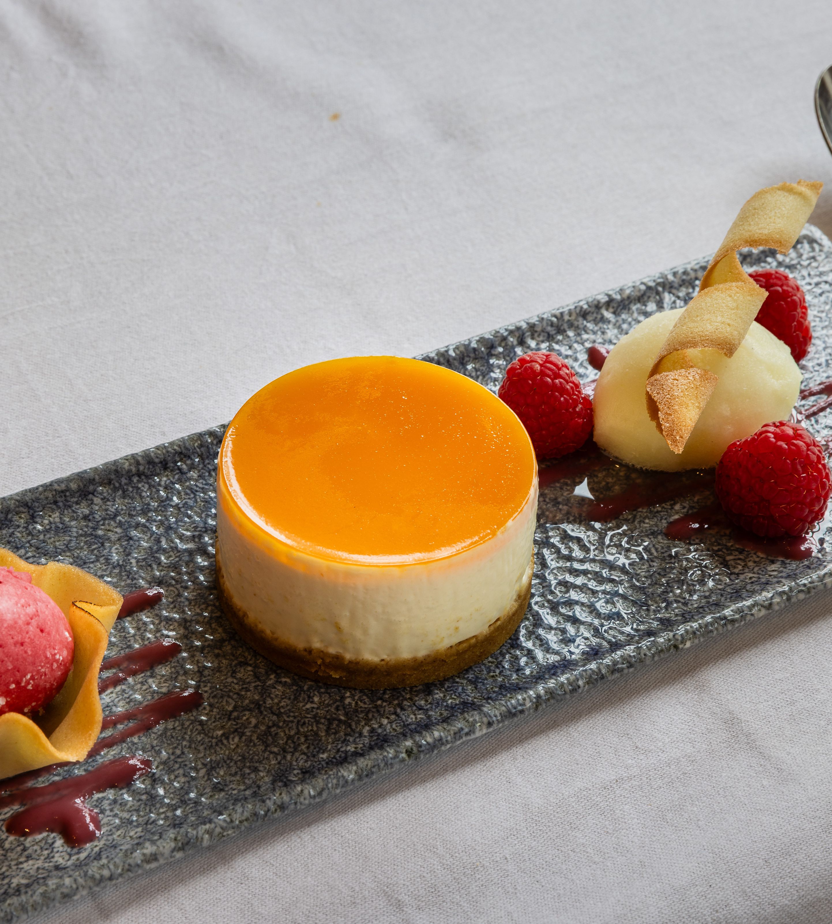 Elegant plated dessert featuring a round cheesecake with orange glaze, raspberry sorbet in a tuile cup, fresh raspberries, and a quenelle of white sorbet garnished with a crisp wafer.