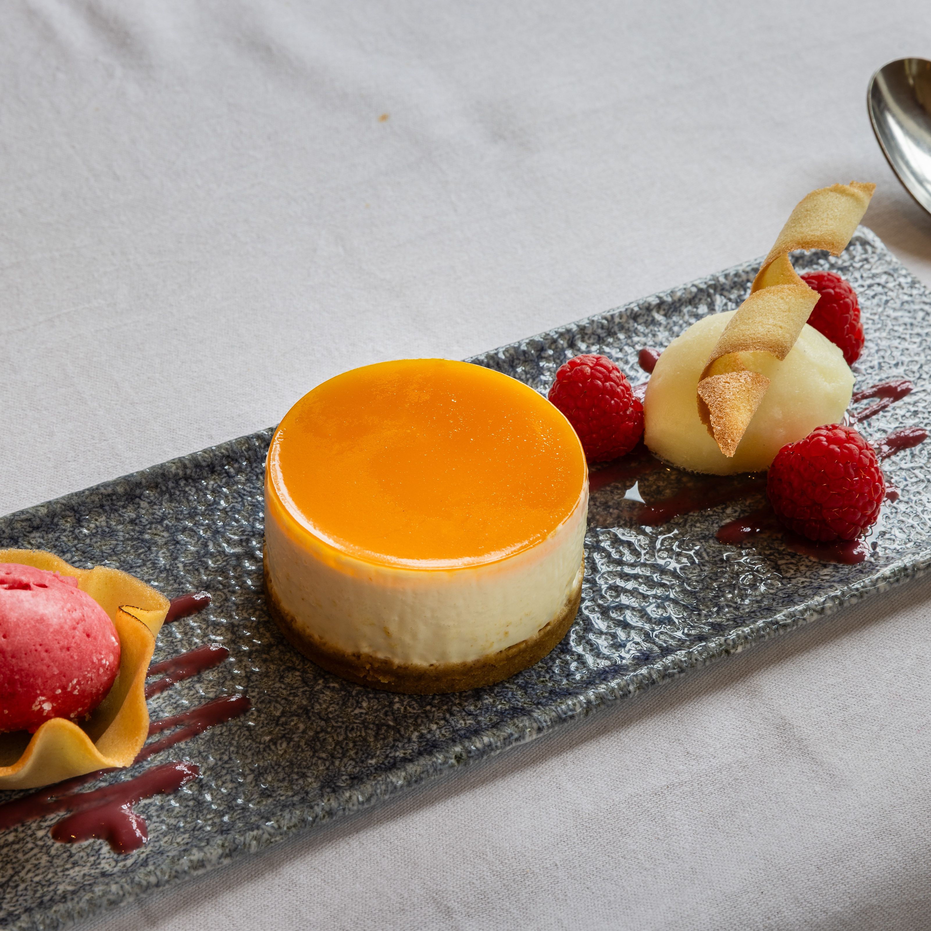 Elegant plated dessert featuring a round cheesecake with orange glaze, raspberry sorbet in a tuile cup, fresh raspberries, and a quenelle of white sorbet garnished with a crisp wafer.