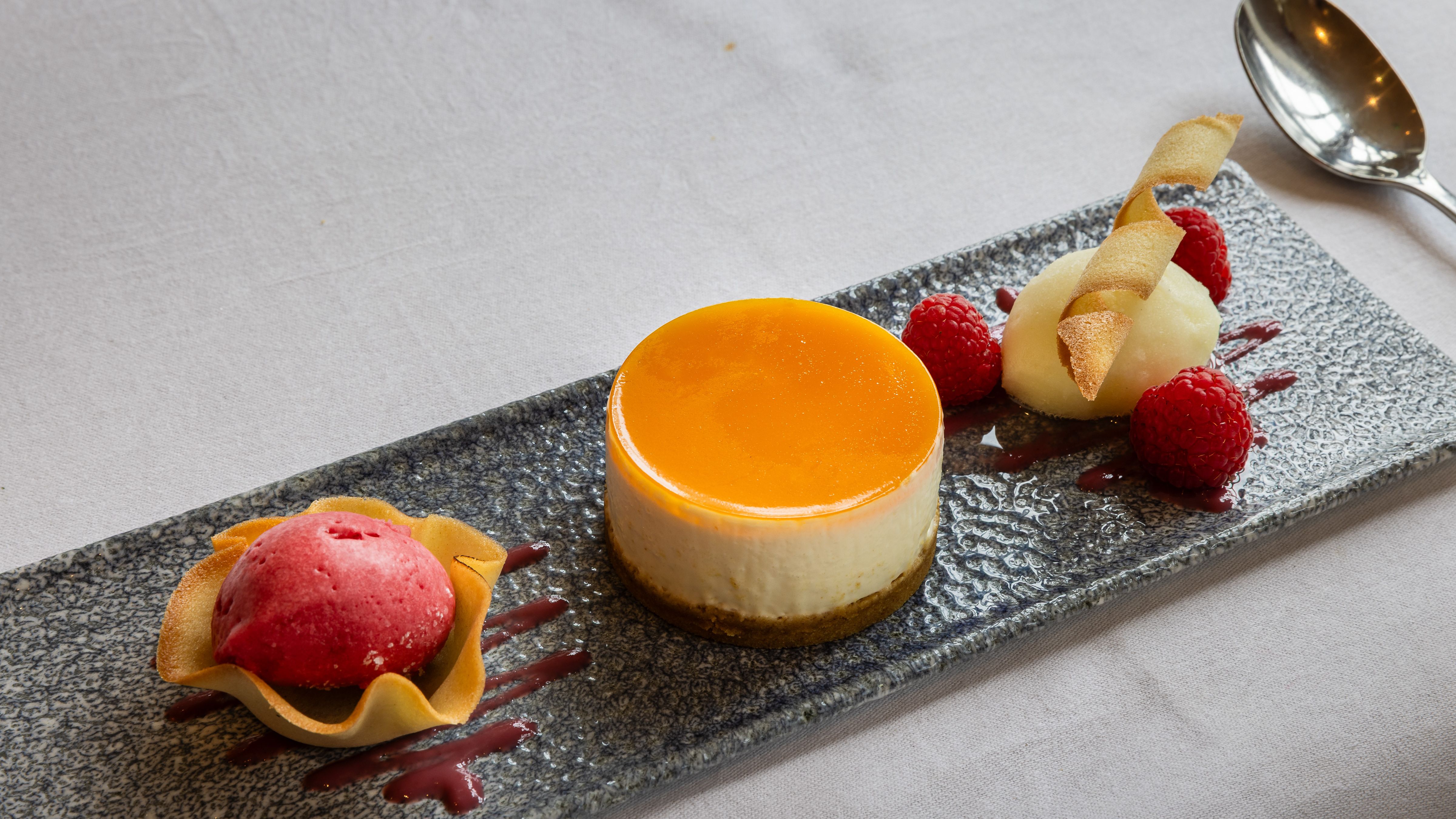 Elegant plated dessert featuring a round cheesecake with orange glaze, raspberry sorbet in a tuile cup, fresh raspberries, and a quenelle of white sorbet garnished with a crisp wafer.