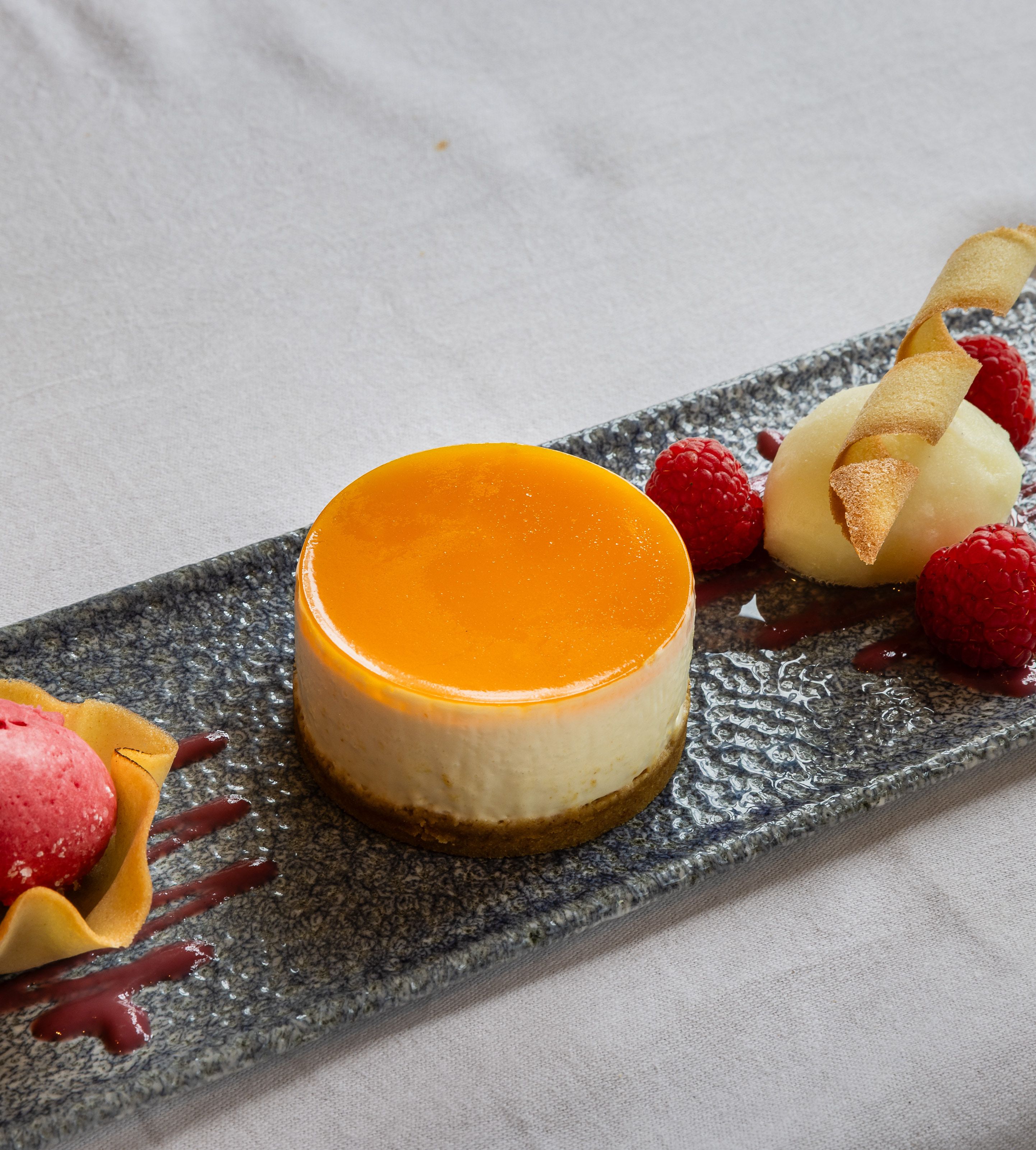 Elegant plated dessert featuring a round cheesecake with orange glaze, raspberry sorbet in a tuile cup, fresh raspberries, and a quenelle of white sorbet garnished with a crisp wafer.