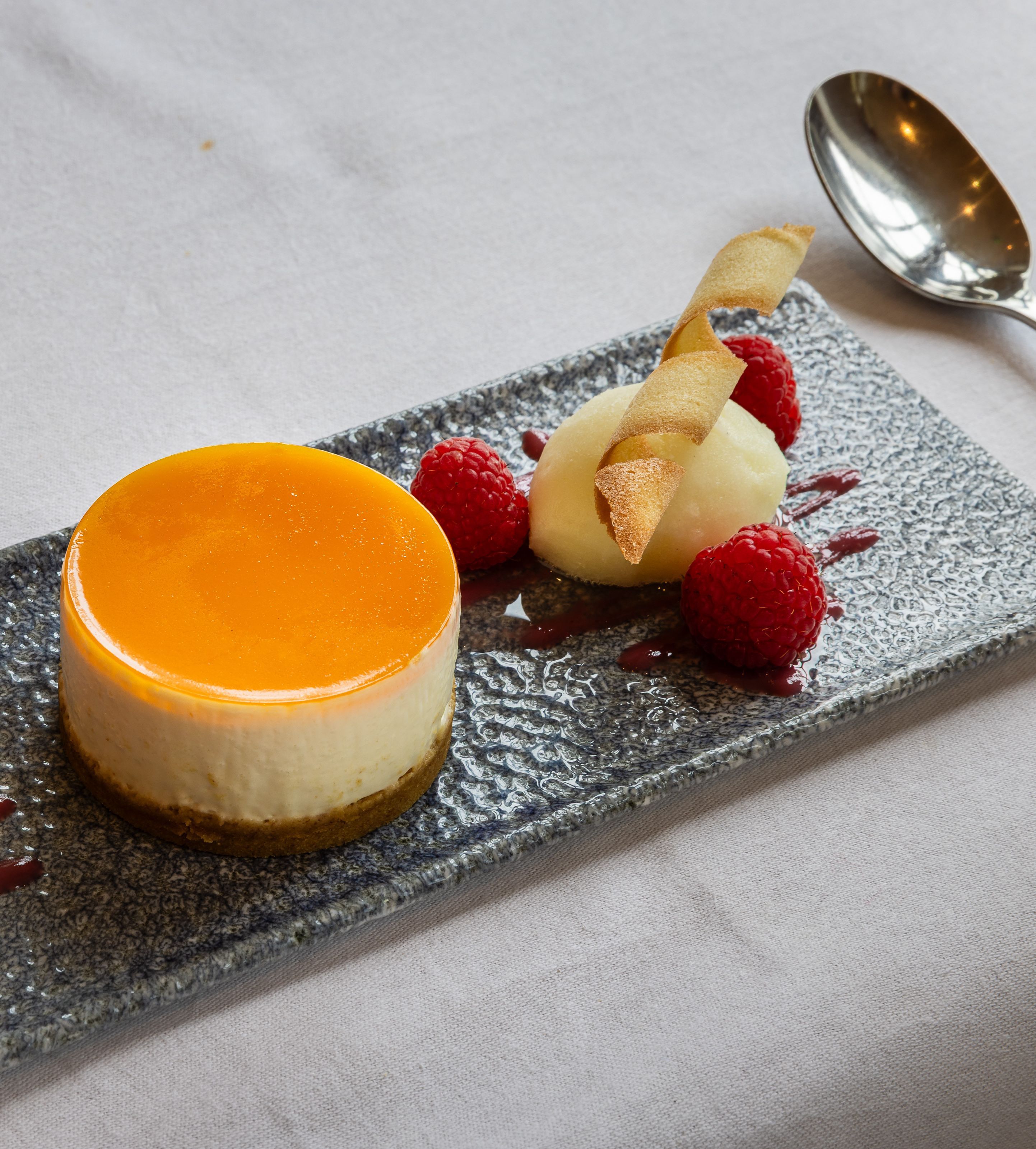 Elegant plated dessert featuring a round cheesecake with orange glaze, raspberry sorbet in a tuile cup, fresh raspberries, and a quenelle of white sorbet garnished with a crisp wafer.