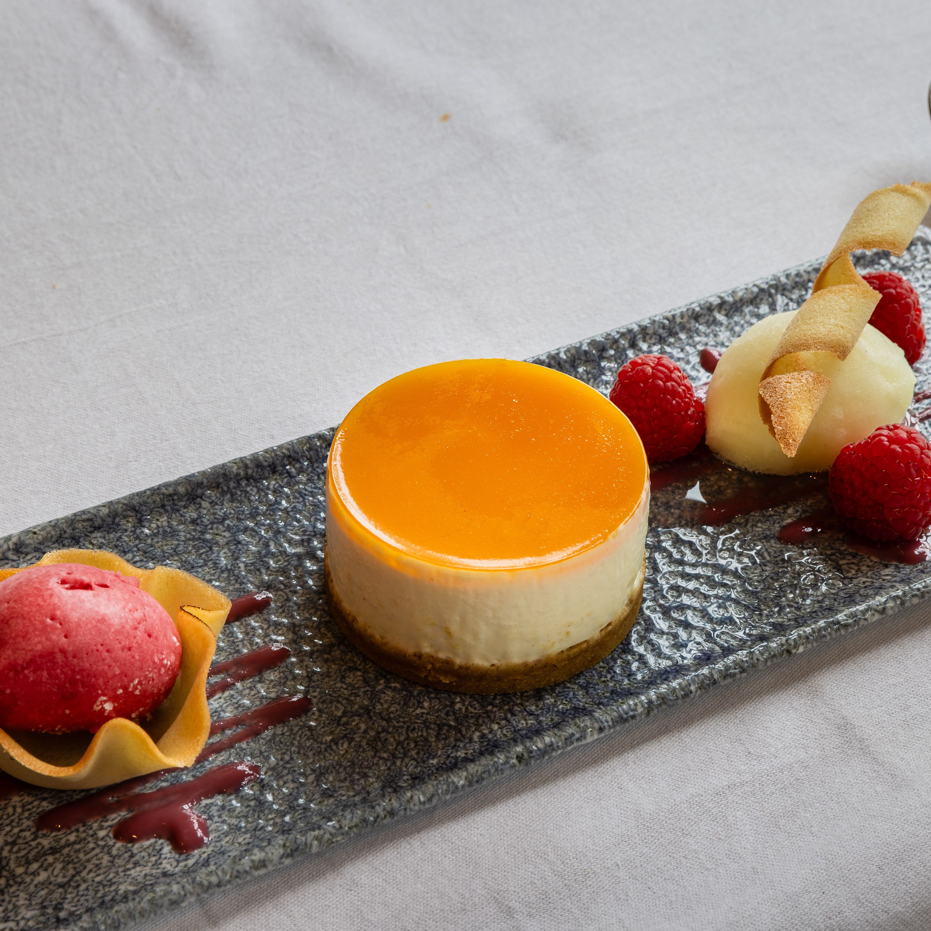 Elegant plated dessert featuring a round cheesecake with orange glaze, raspberry sorbet in a tuile cup, fresh raspberries, and a quenelle of white sorbet garnished with a crisp wafer.