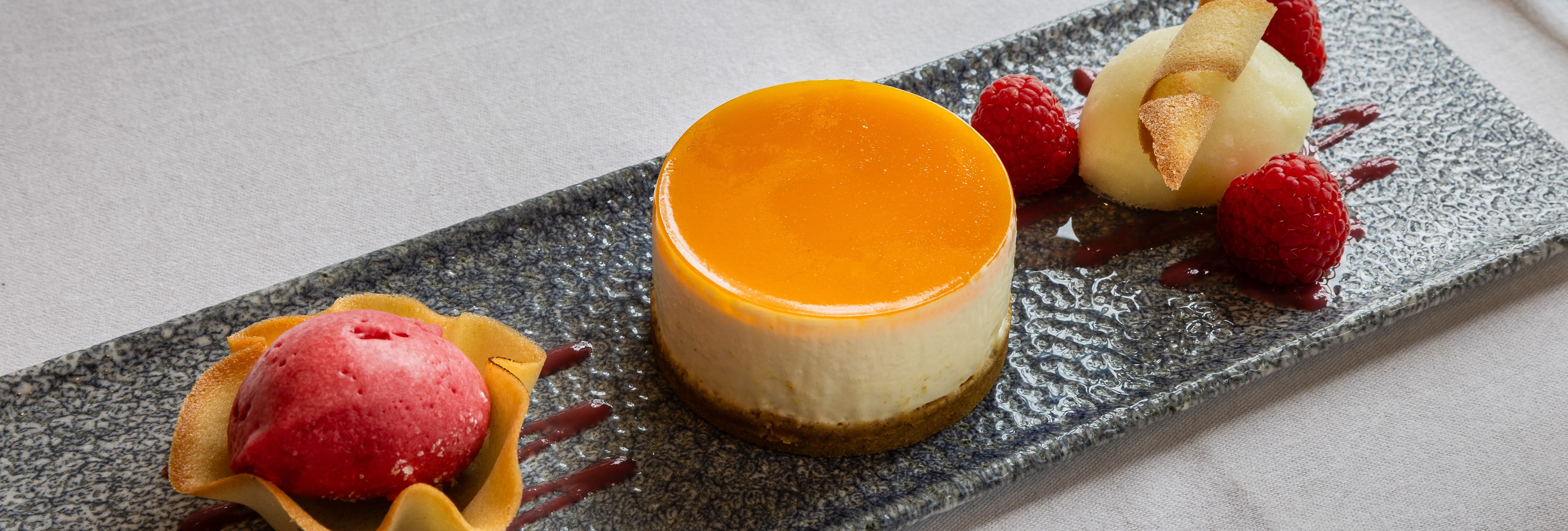 Elegant plated dessert featuring a round cheesecake with orange glaze, raspberry sorbet in a tuile cup, fresh raspberries, and a quenelle of white sorbet garnished with a crisp wafer.