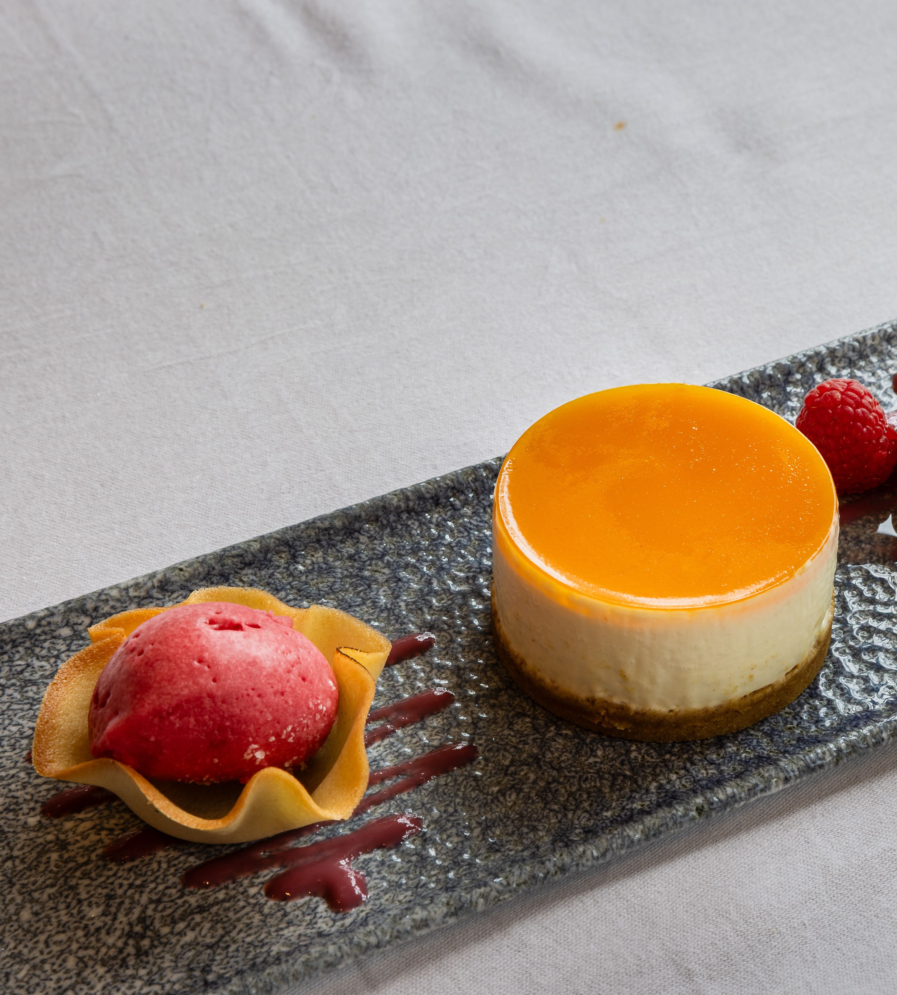 Elegant plated dessert featuring a round cheesecake with orange glaze, raspberry sorbet in a tuile cup, fresh raspberries, and a quenelle of white sorbet garnished with a crisp wafer.