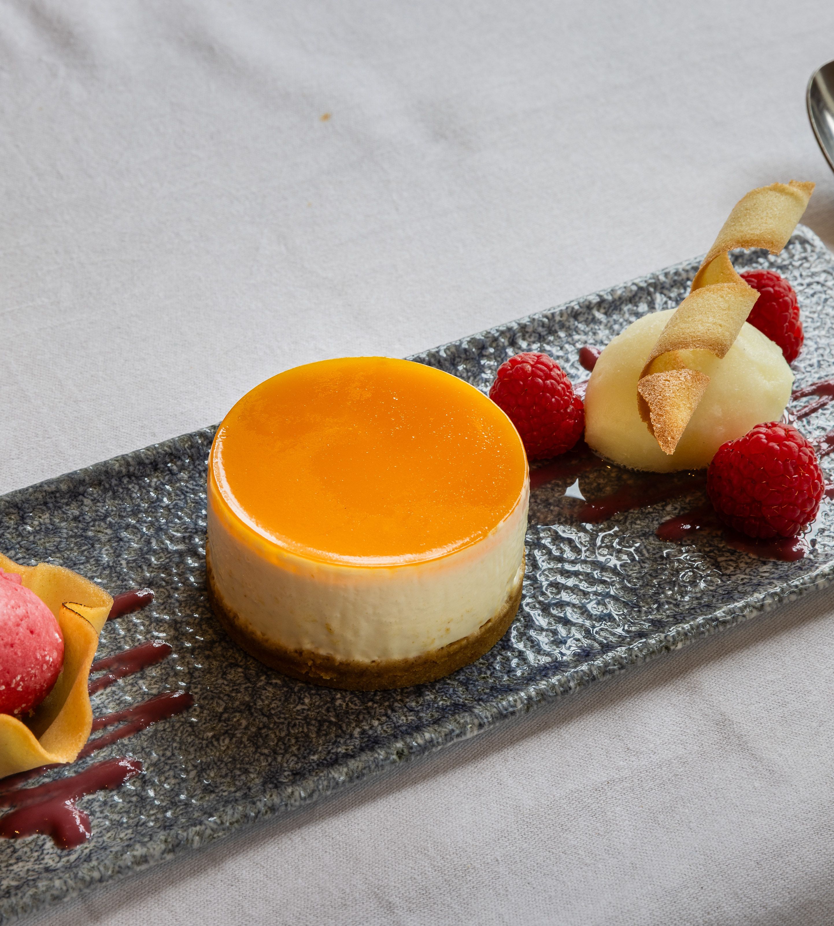 Elegant plated dessert featuring a round cheesecake with orange glaze, raspberry sorbet in a tuile cup, fresh raspberries, and a quenelle of white sorbet garnished with a crisp wafer.