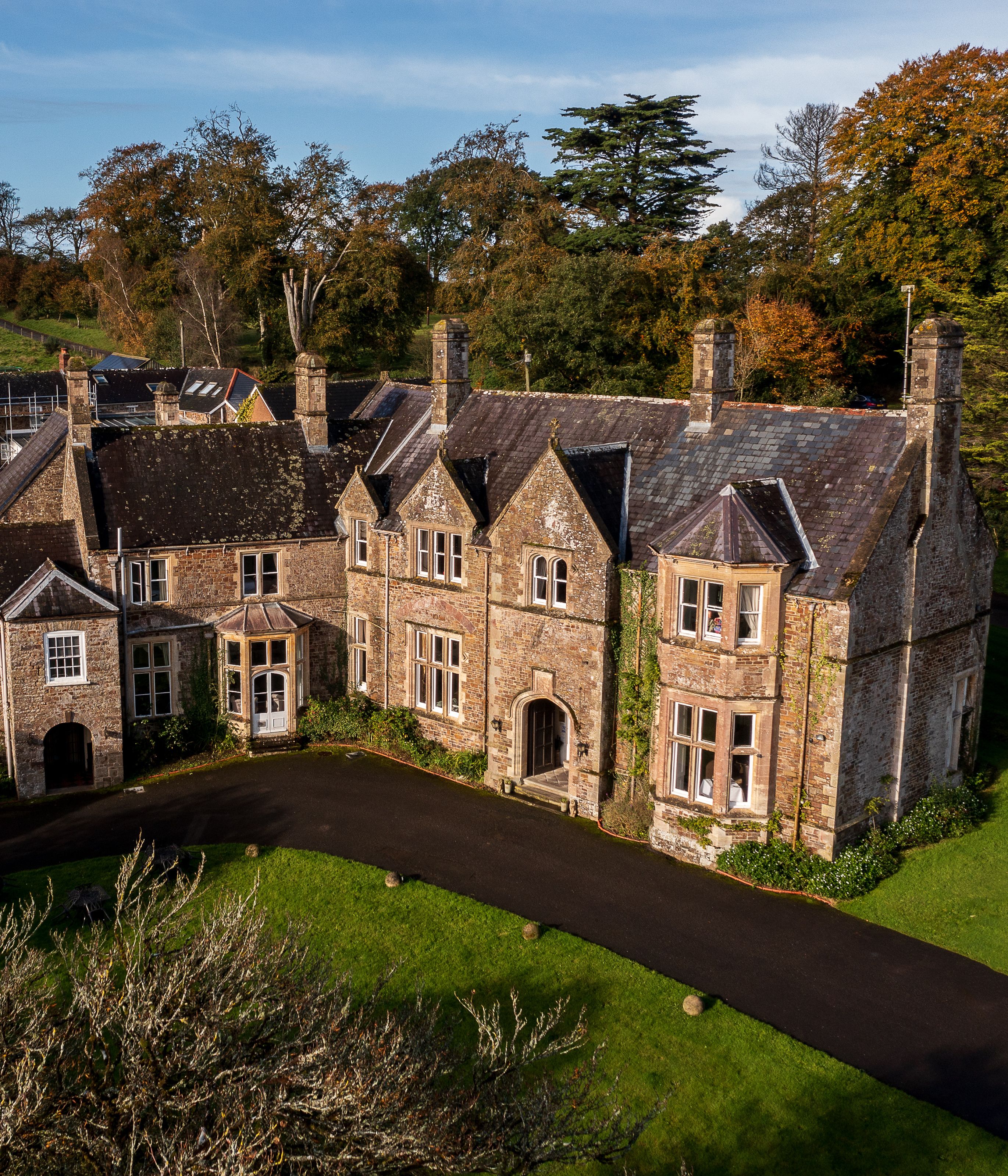 Aerial view of a large, historic stone manor house surrounded by gardens and countryside.