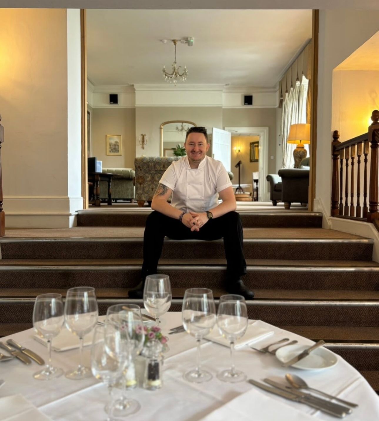 A chef sitting on stairs in a refined dining area with a table set for a meal in the foreground.