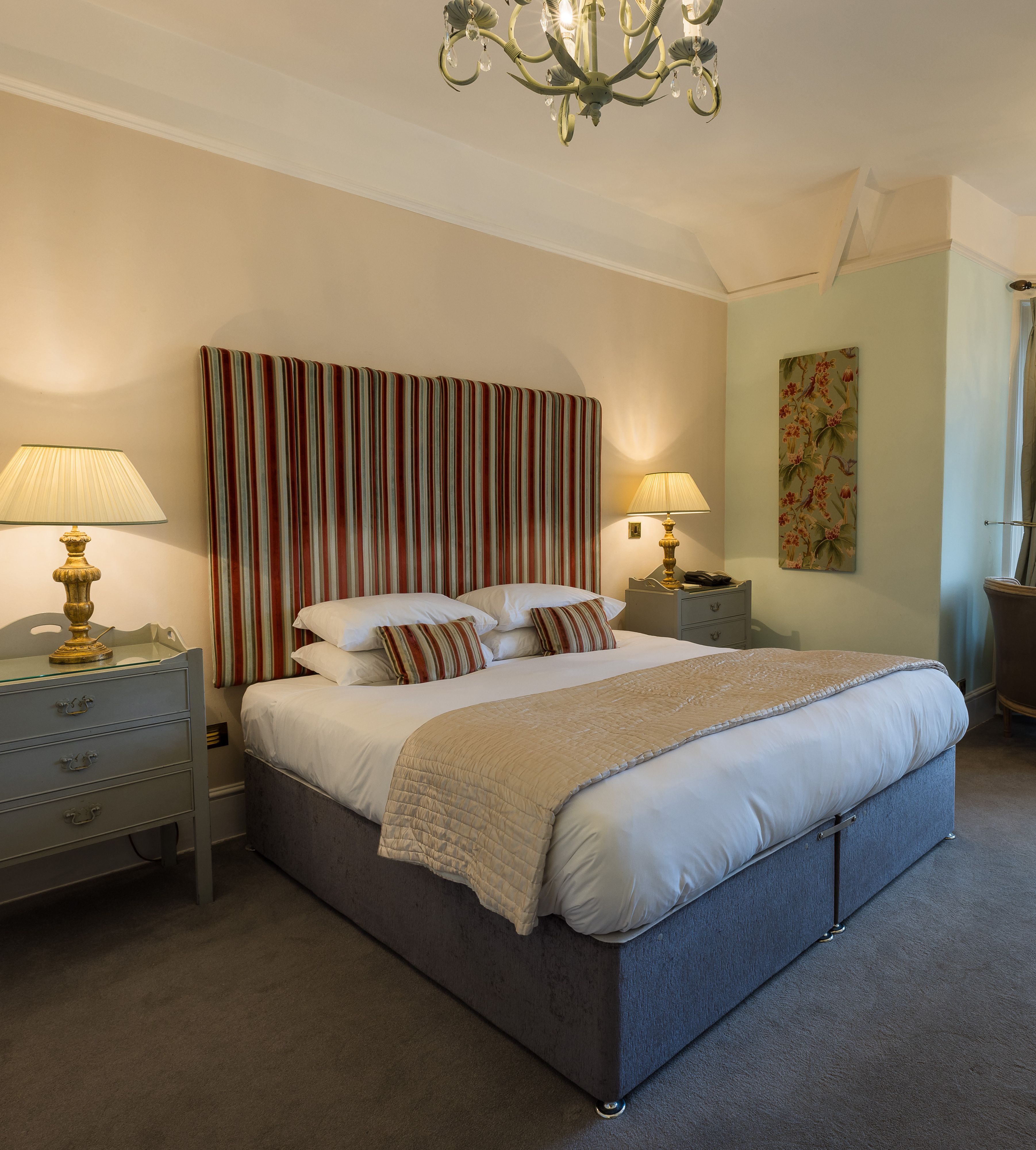 Elegant hotel bedroom with a large double bed, striped headboard, bedside tables with lamps, armchairs, and tall windows.