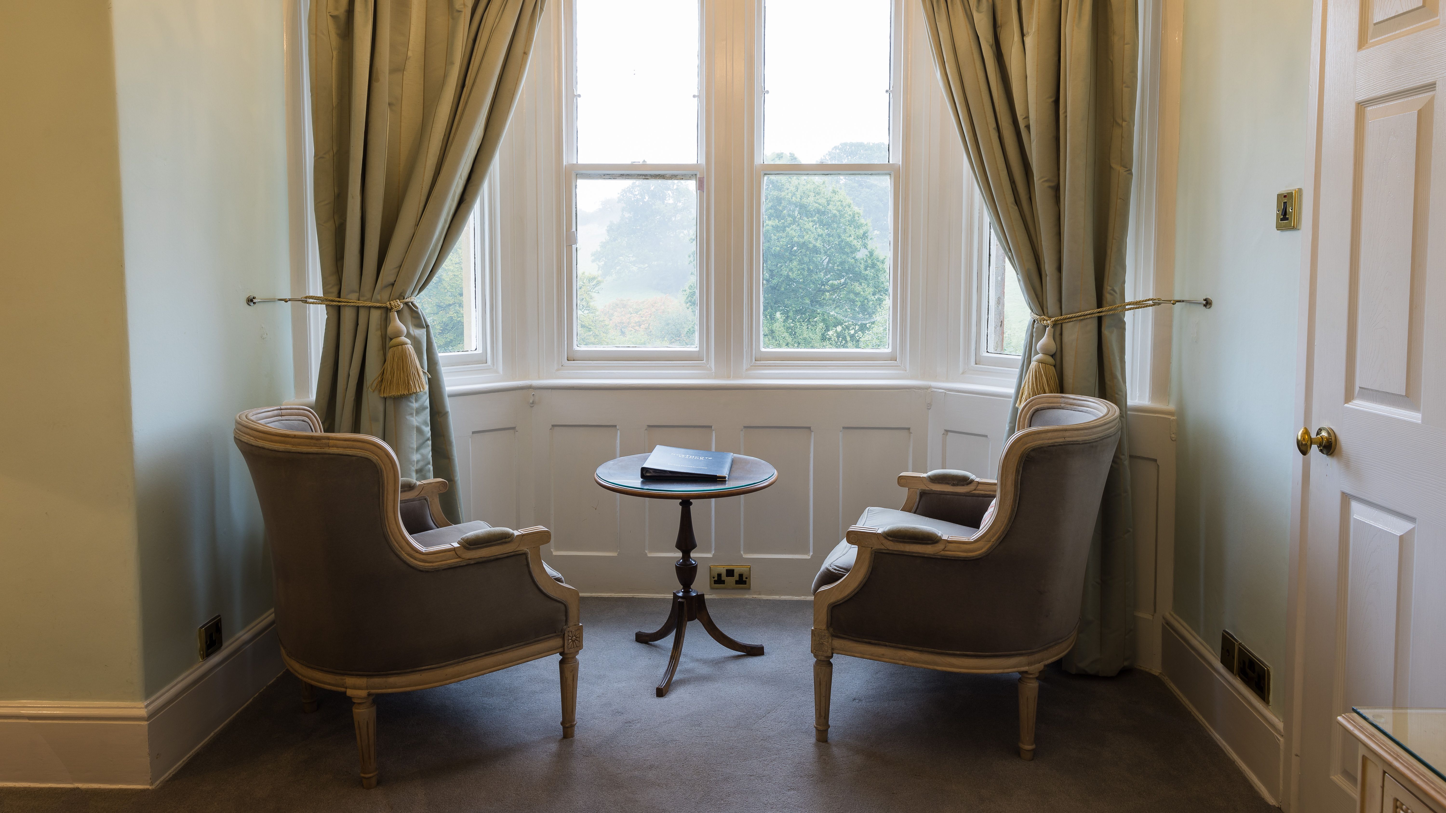 Elegant sitting area with two armchairs, a small round table, and large bay windows with curtains.