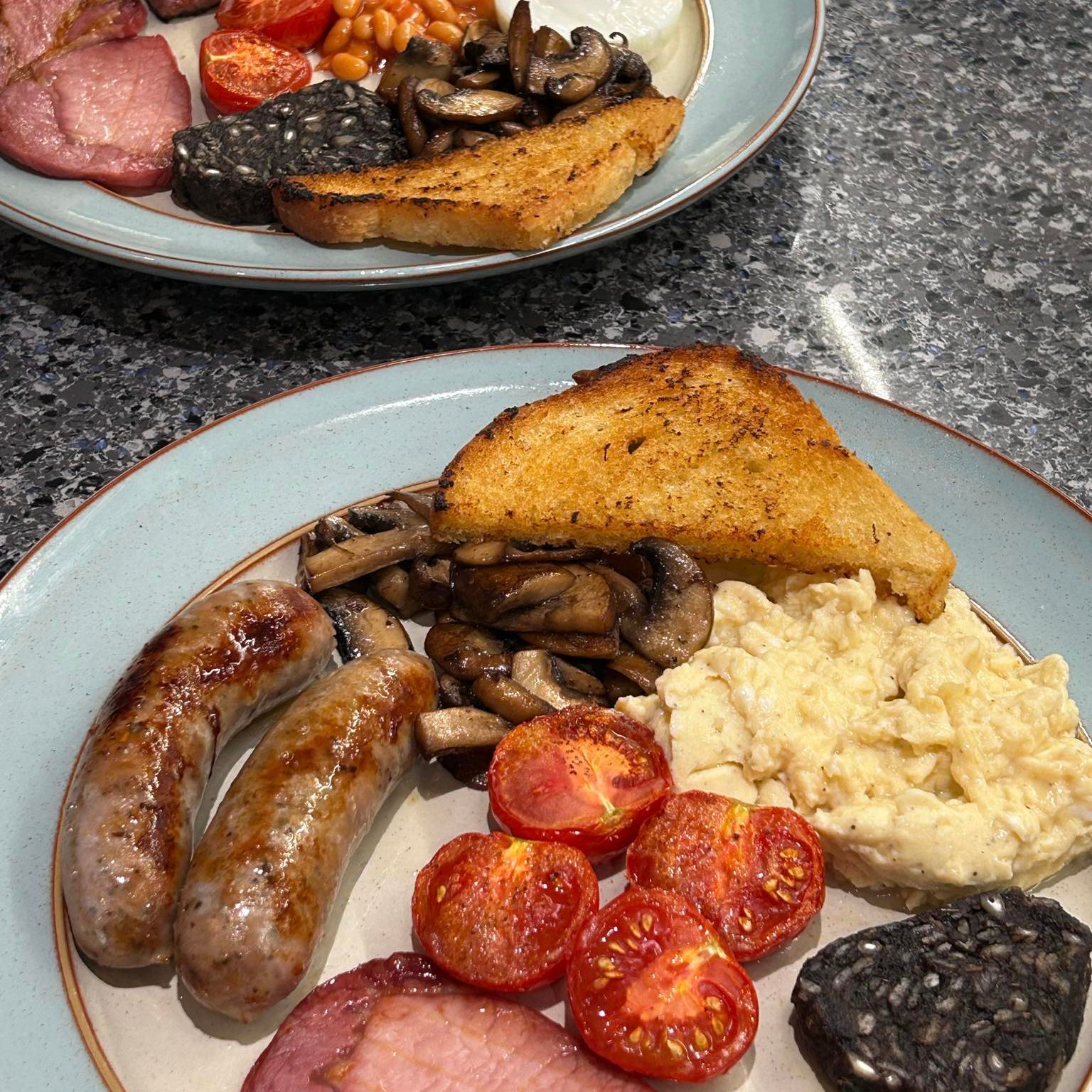 Two plates of full english breakfasts with eggs, sausages, black pudding, bacon,tomatoes, mushrooms and fried bread