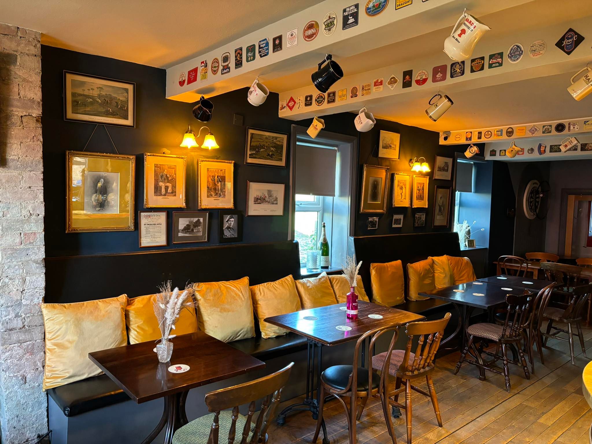 Cozy pub interior with framed pictures, yellow cushions, and hanging mugs