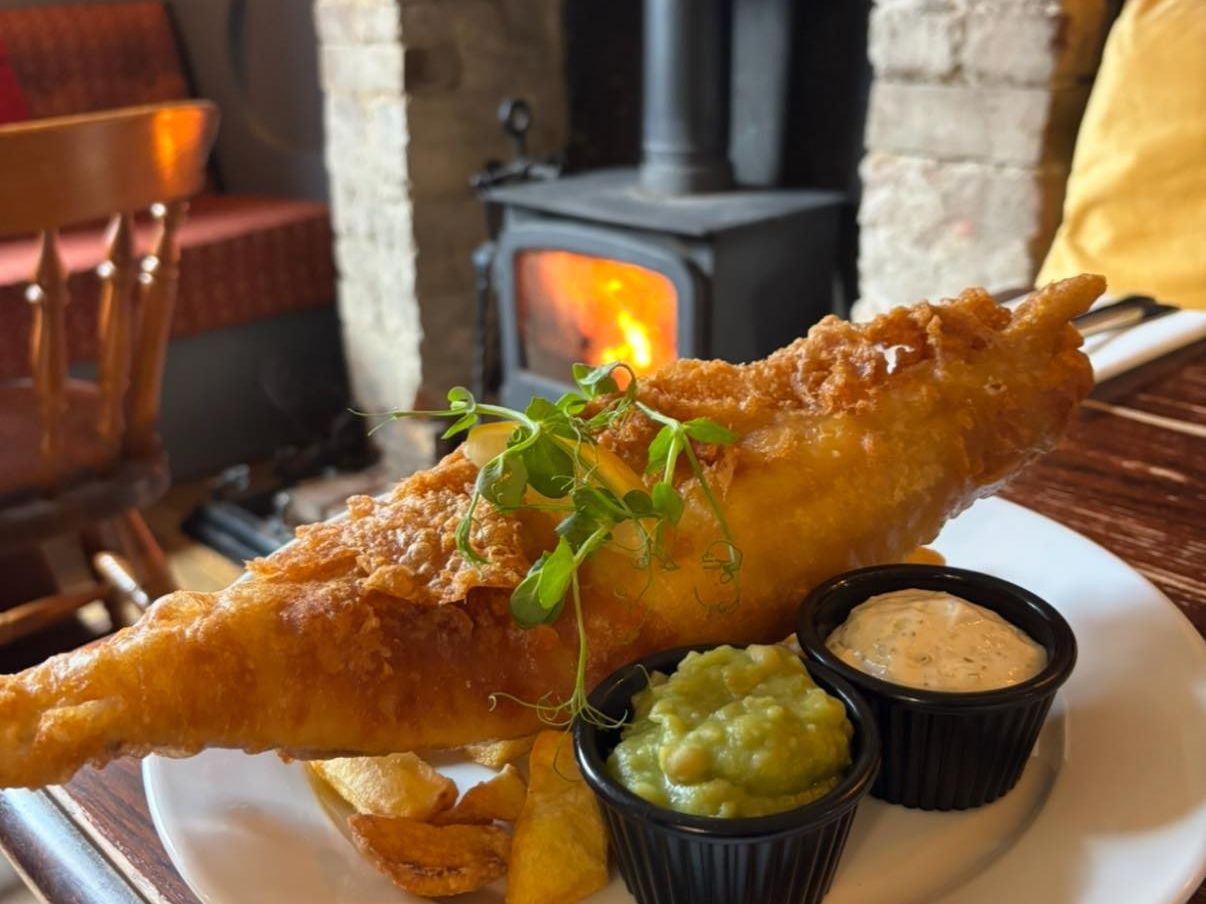 Plate of fish and chips with mushy peas and tartar sauce on a wooden table in front of a lit fireplace.