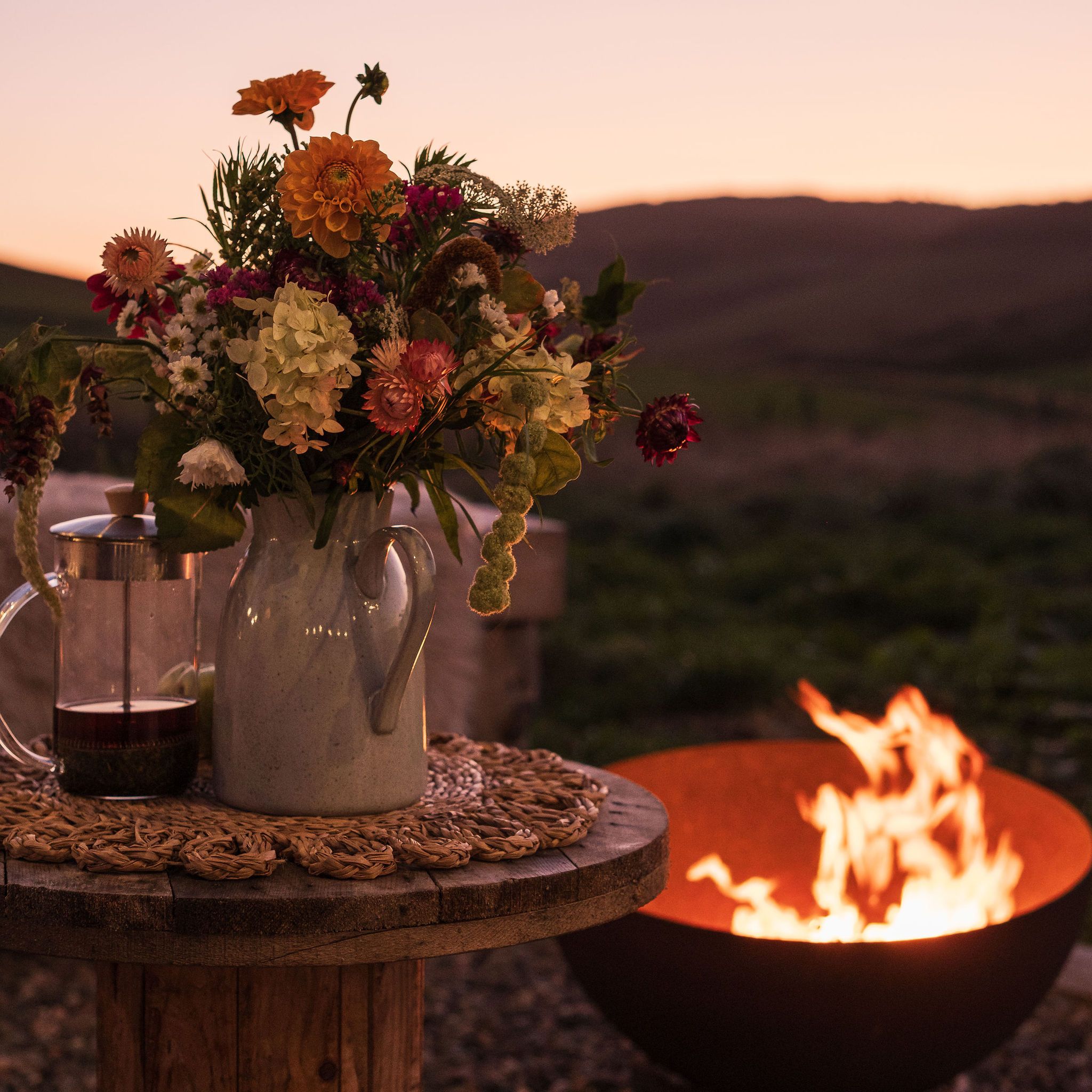 Wildflower bouquet in a pitcher on a rustic wooden table beside an outdoor fire pit at sunset